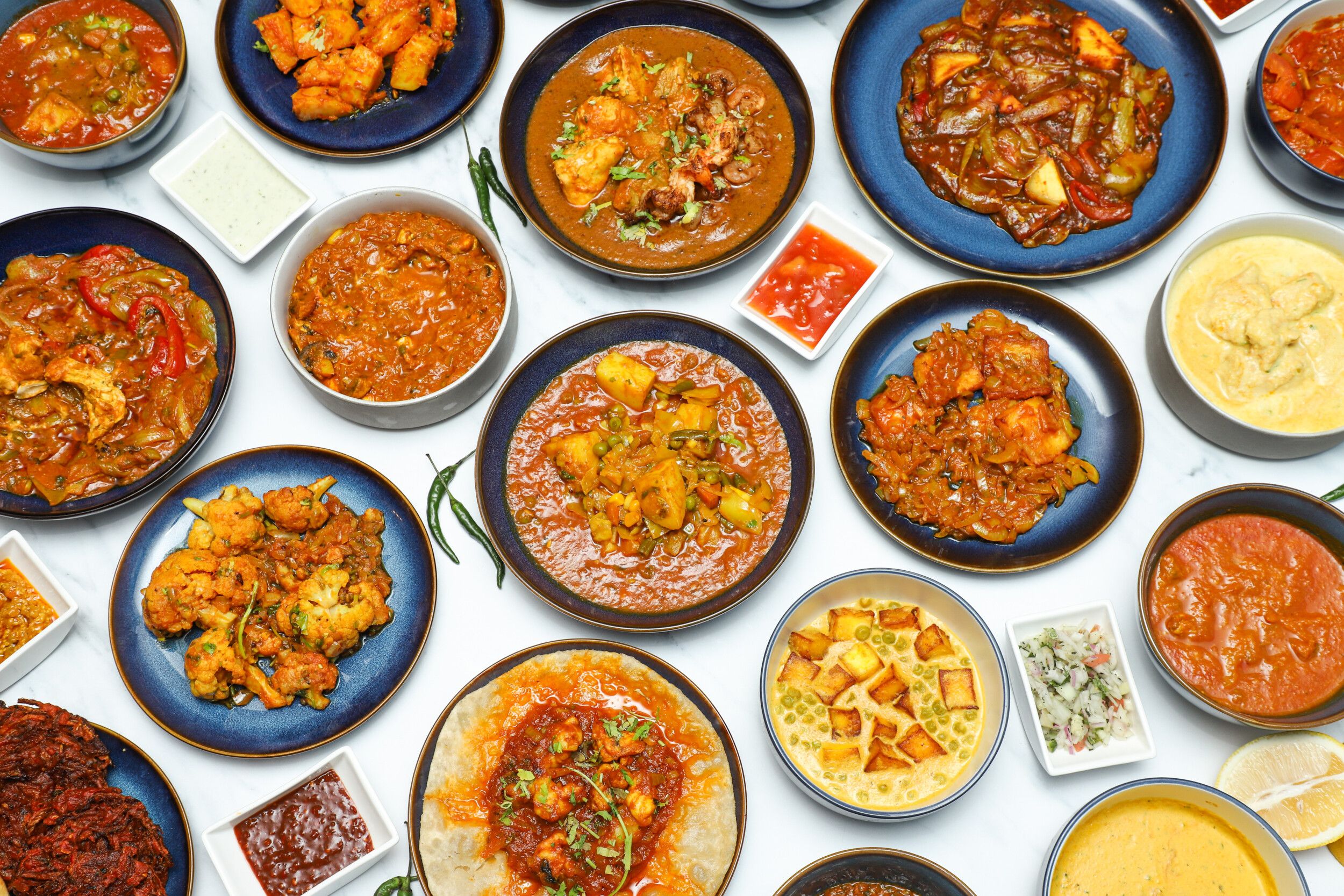 An overhead shot of various Indian curries, sides, and accompaniments in bowls and plates on a white background, including chutneys, cauliflower, paneer, and