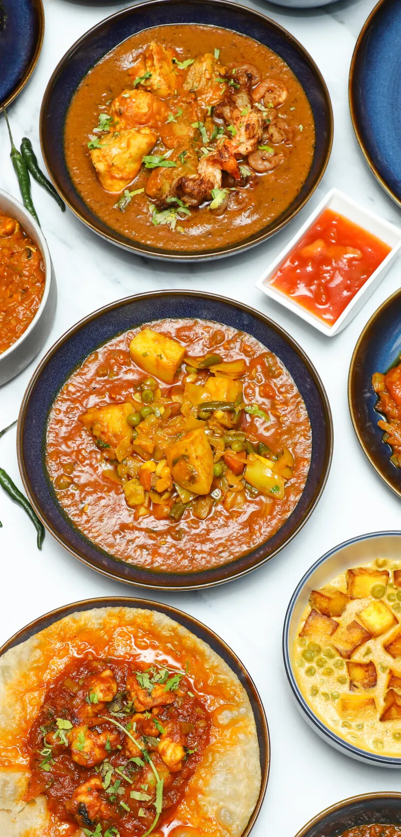 An overhead shot of various Indian curries, sides, and accompaniments in bowls and plates on a white background, including chutneys, cauliflower, paneer, and