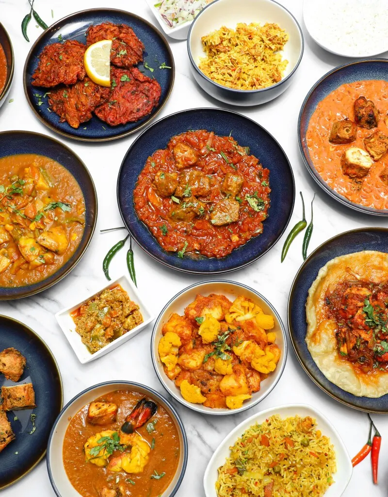 An overhead shot of various Indian curries, rice, appetizers, and breads arranged on a white table.