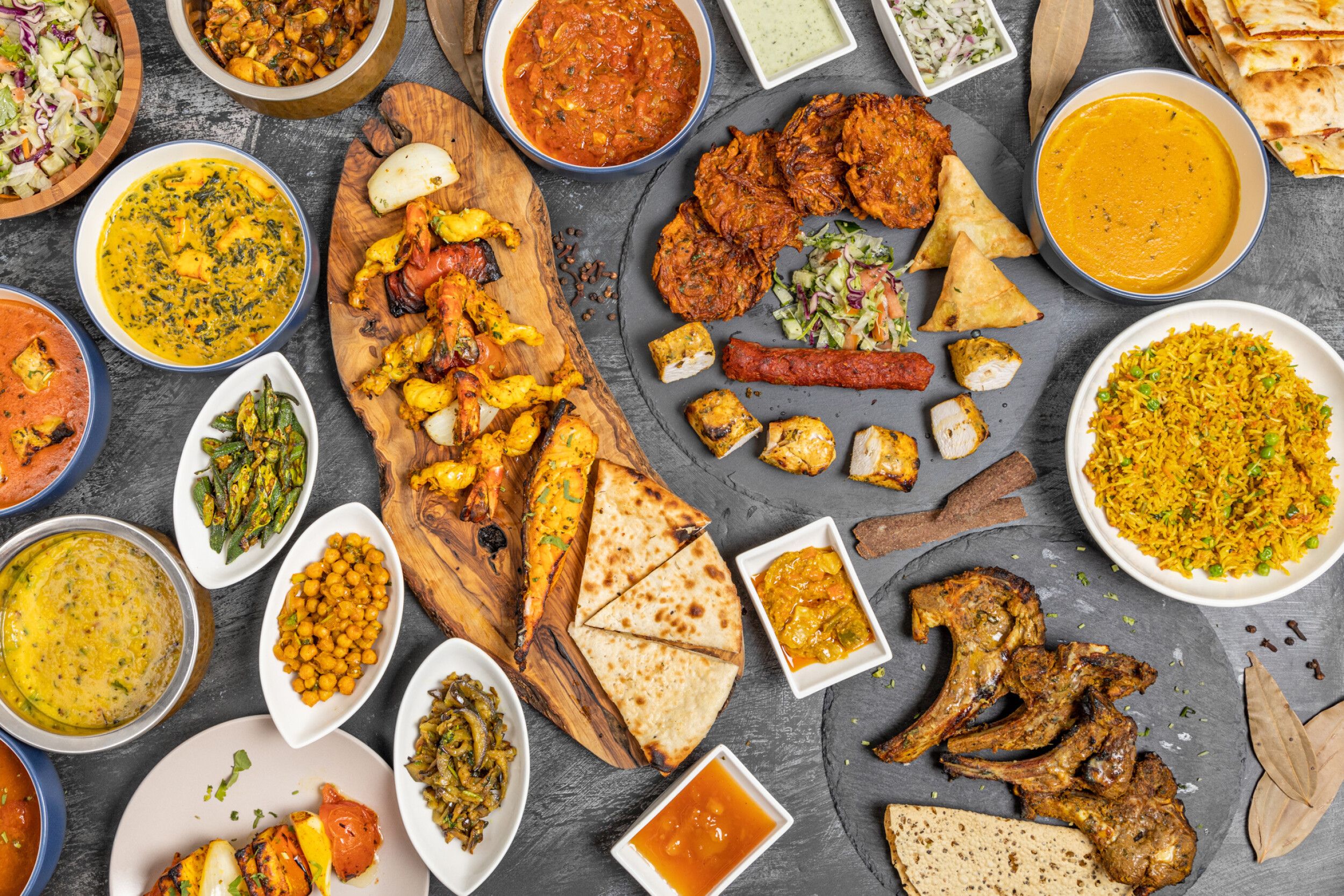 An overhead shot of a large spread of various Indian dishes on a dark grey surface.