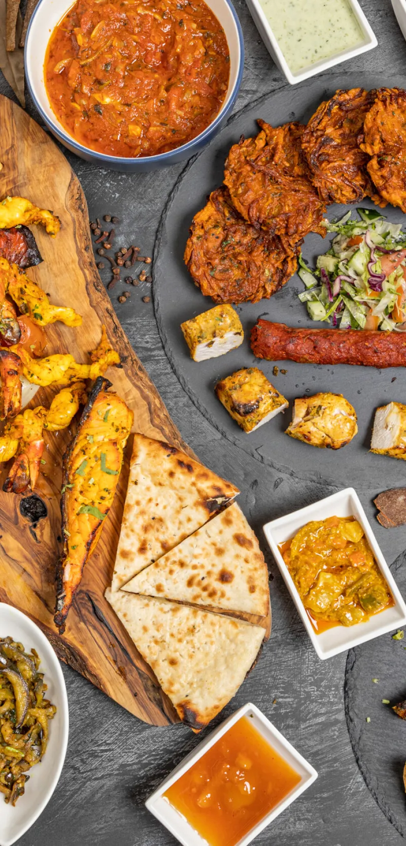 An overhead shot of a large spread of various Indian dishes on a dark grey surface.