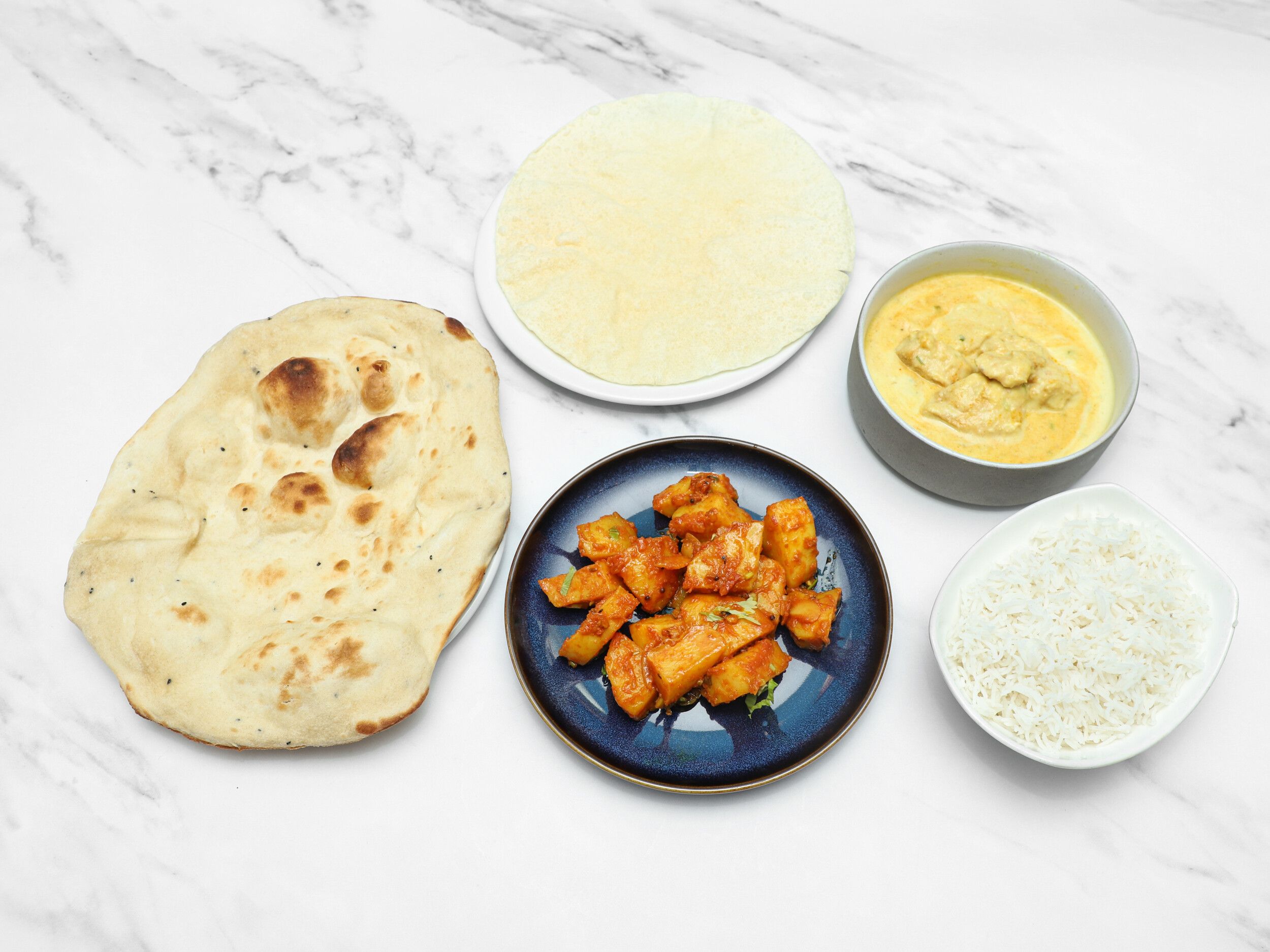 Indian naan bread, white rice, chicken curry, and aloo gobi on white marble surface