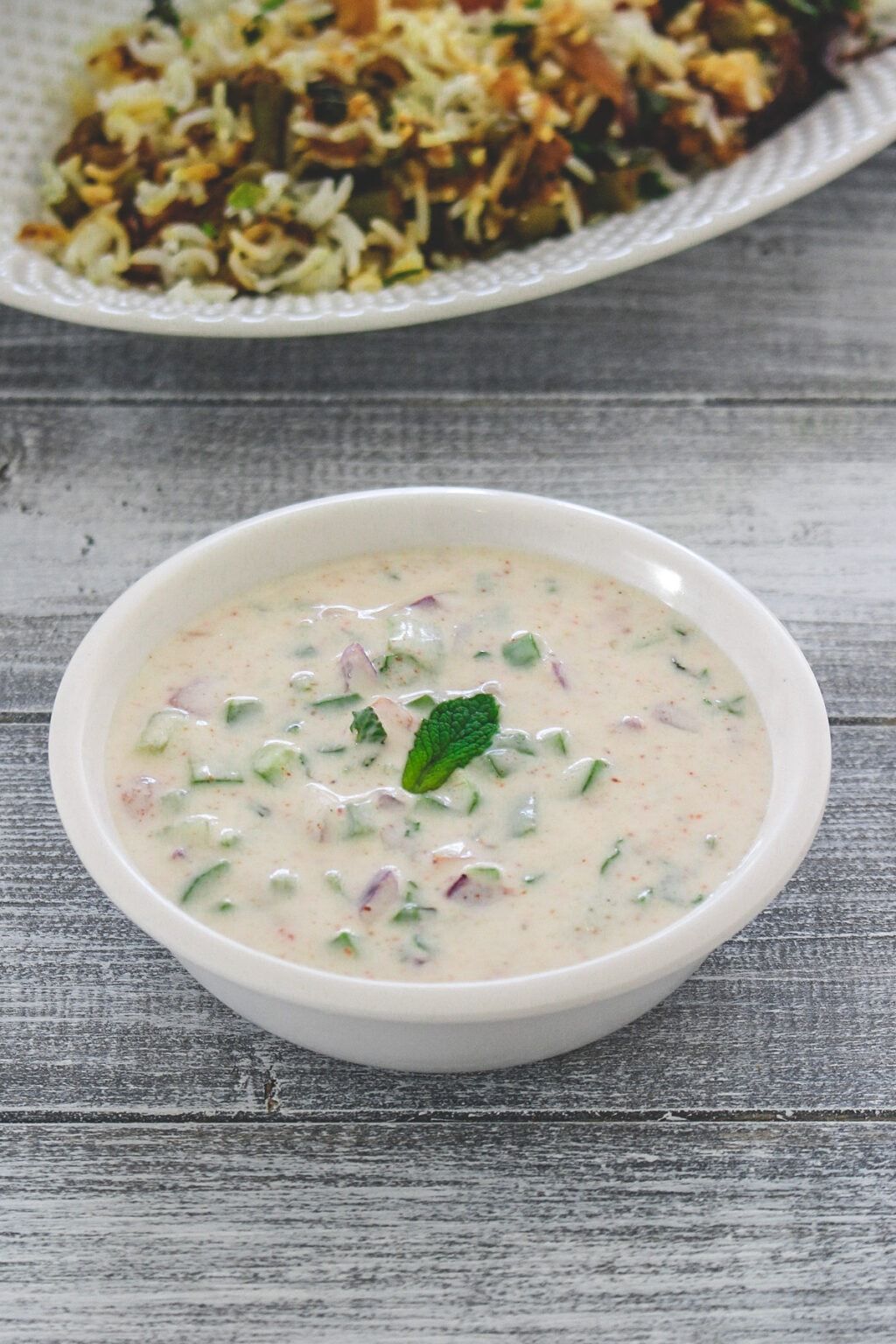 A white bowl of raita with cucumber and red onion, garnished with mint, next to biryani.