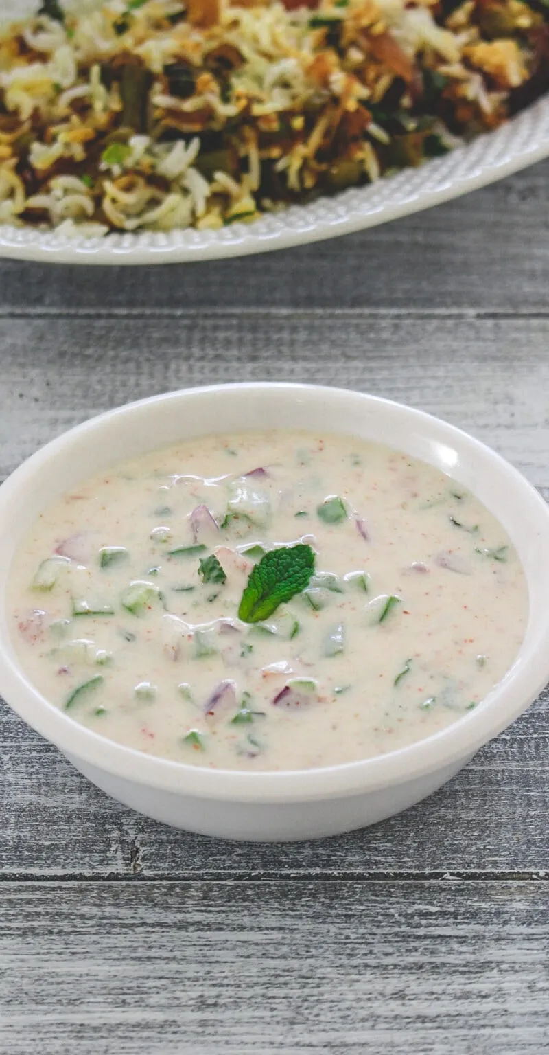 A white bowl of raita with cucumber and red onion, garnished with mint, next to biryani.