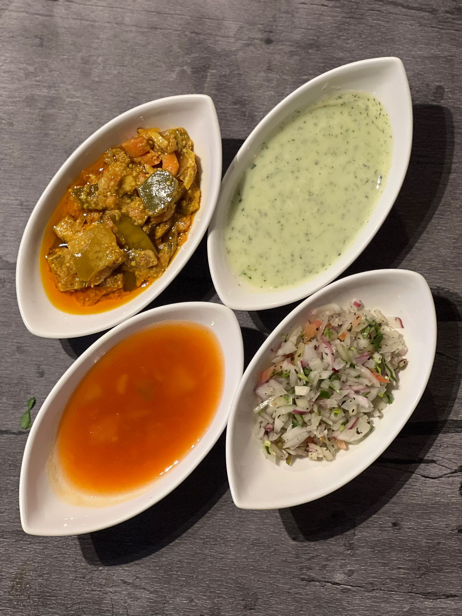 Four leaf-shaped white bowls with orange curry, green raita, red sauce, and onion salad.