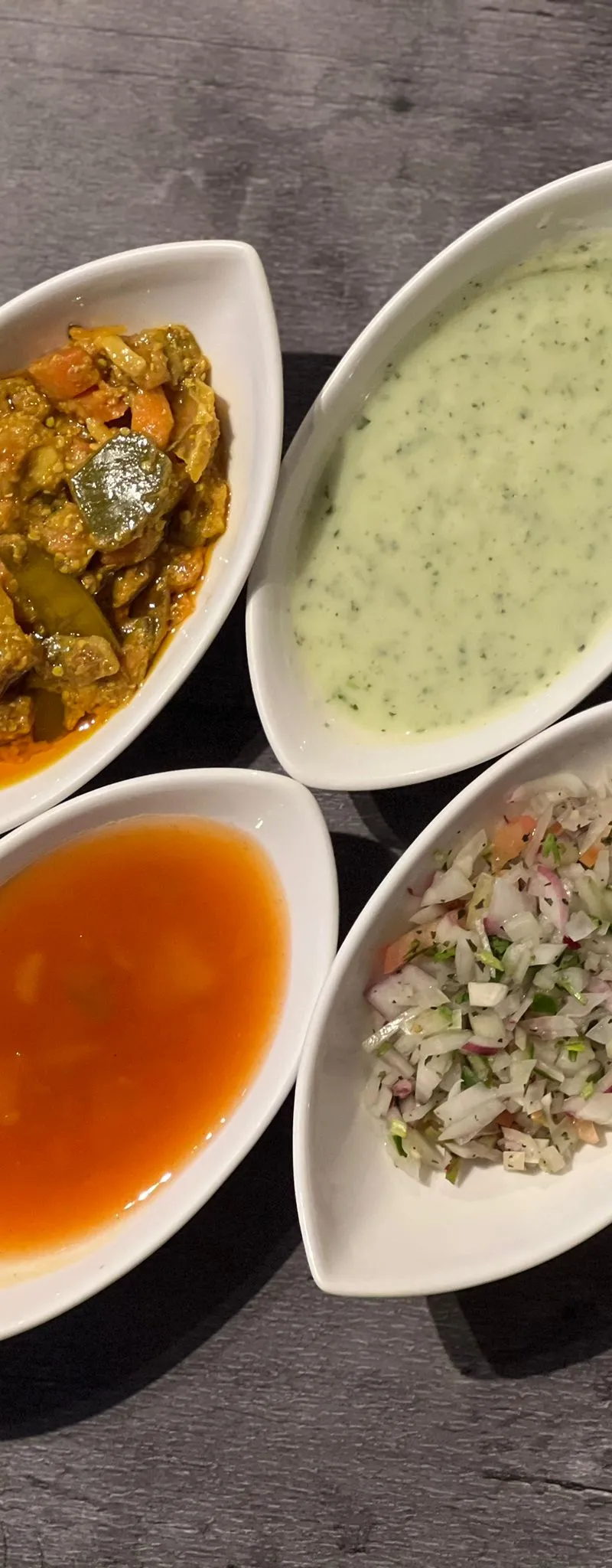 Four leaf-shaped white bowls with orange curry, green raita, red sauce, and onion salad.