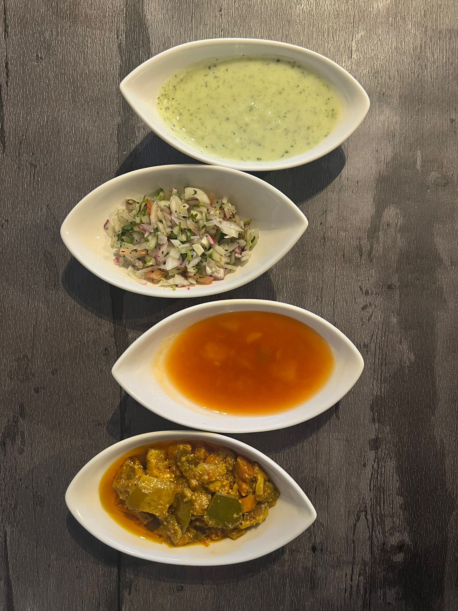 Four small white oval dishes on a dark wooden surface, containing raita, salad, chutney, and pickle.