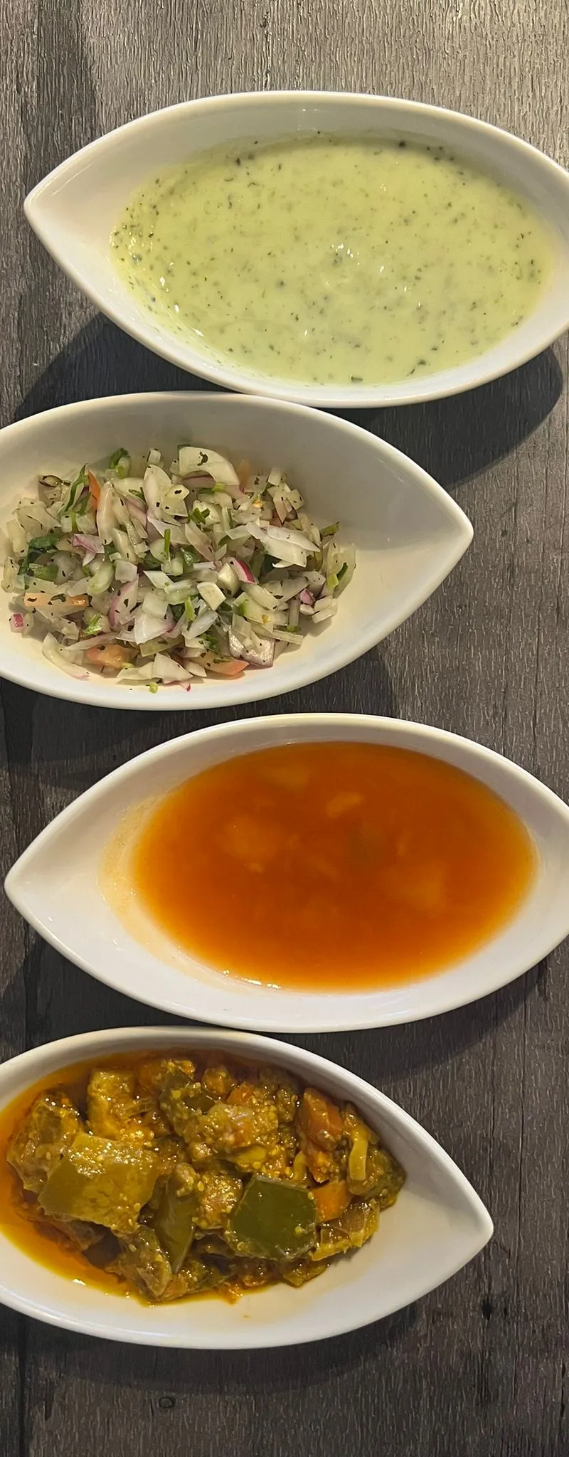 Four small white oval dishes on a dark wooden surface, containing raita, salad, chutney, and pickle.