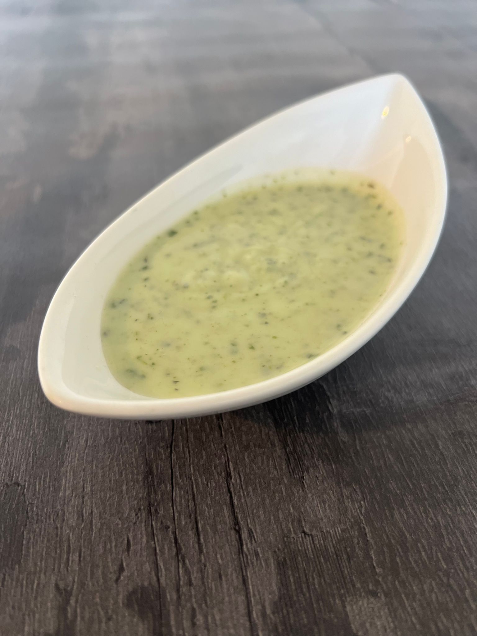 Light green mint sauce with visible herb flecks in an elegant white, boat-shaped dish on a dark wooden surface.