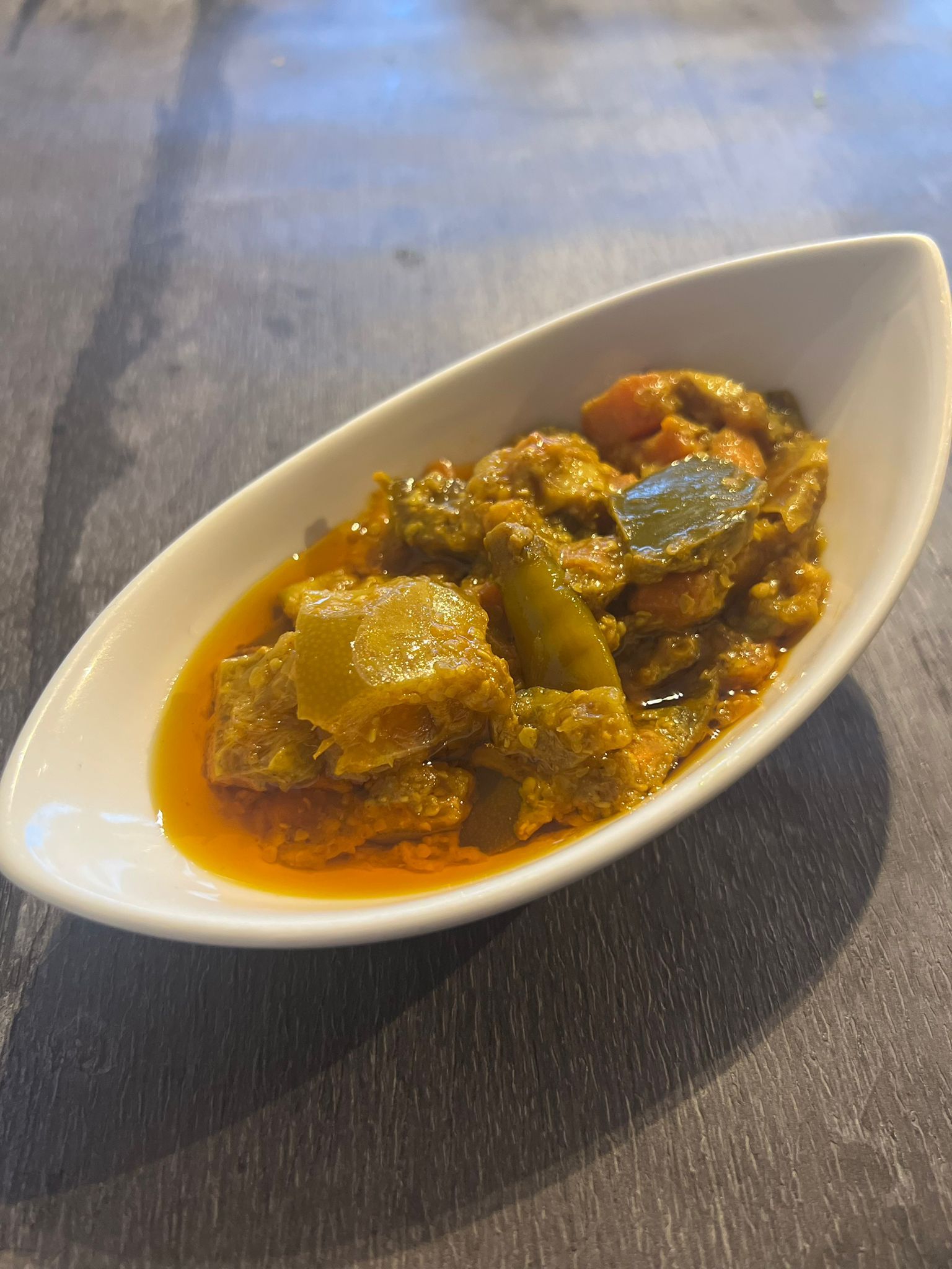 Indian pickle with green, orange, and yellow pieces in a white boat-shaped dish on a dark wooden table.