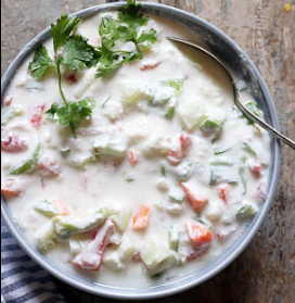 A pale blue bowl of raita with diced cucumber, tomato, and fresh coriander leaves