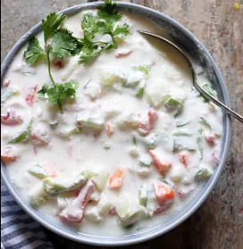 A pale blue bowl of raita with diced cucumber, tomato, and fresh coriander leaves