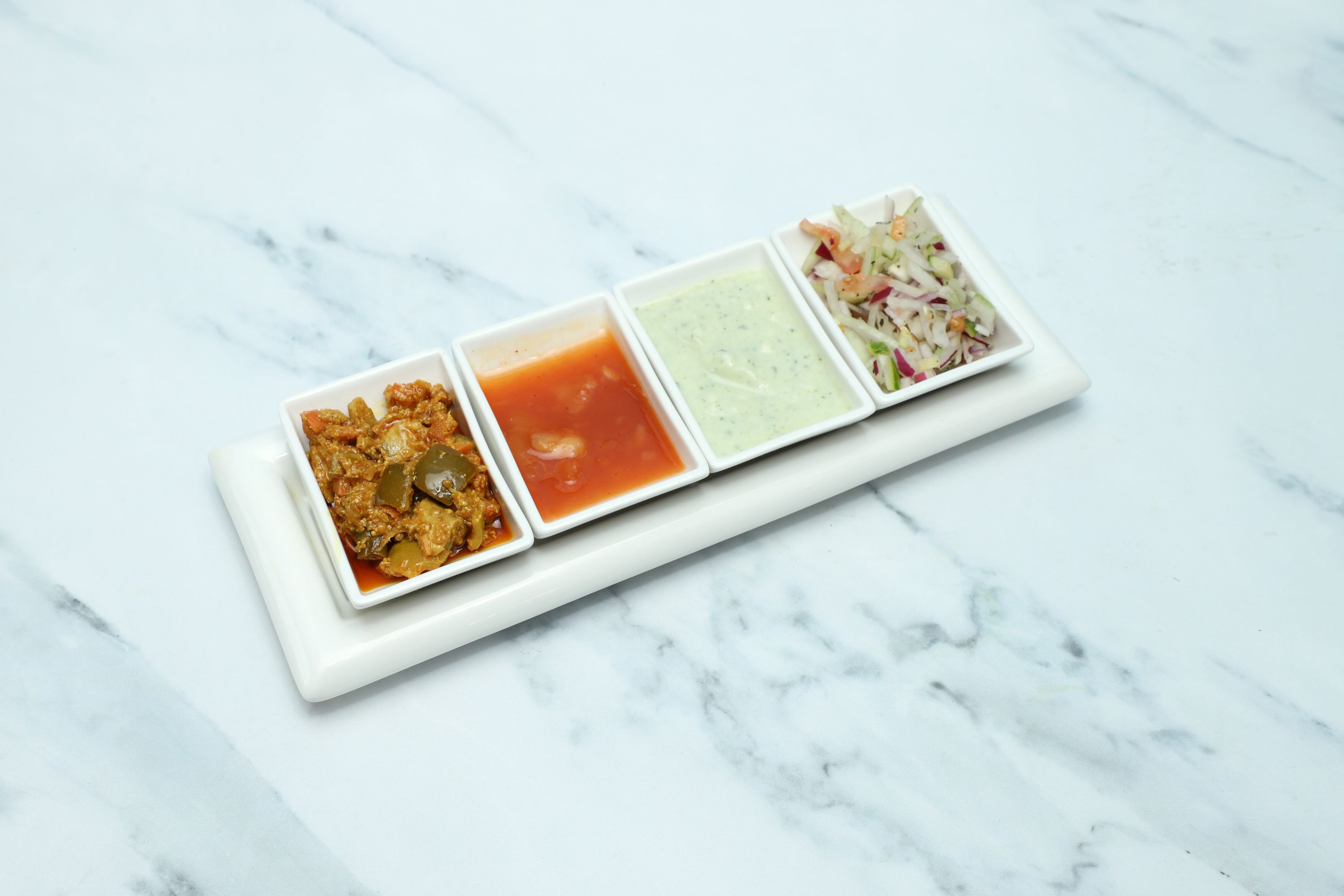 A white rectangular tray holds four small white containers with various condiments on a marble surface.