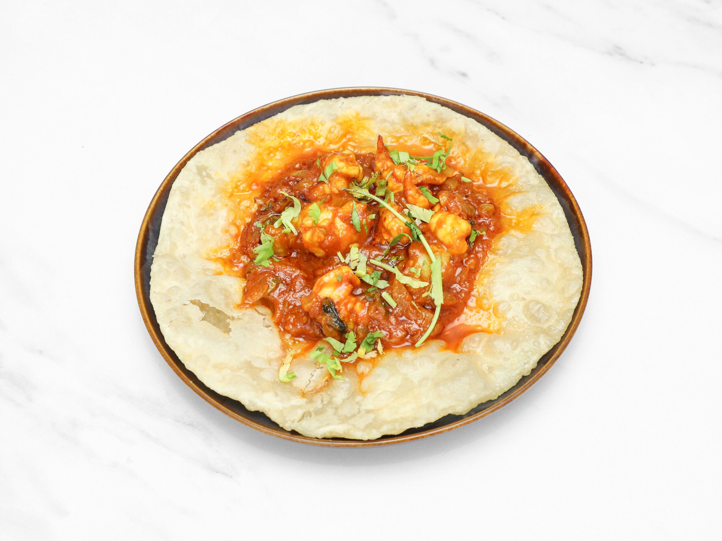 Top-down view of a golden fried puri topped with saucy prawn curry and fresh coriander.