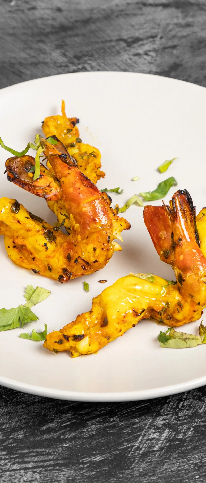 Grilled prawns on skewers, garnished with green herbs, served on a round white plate.