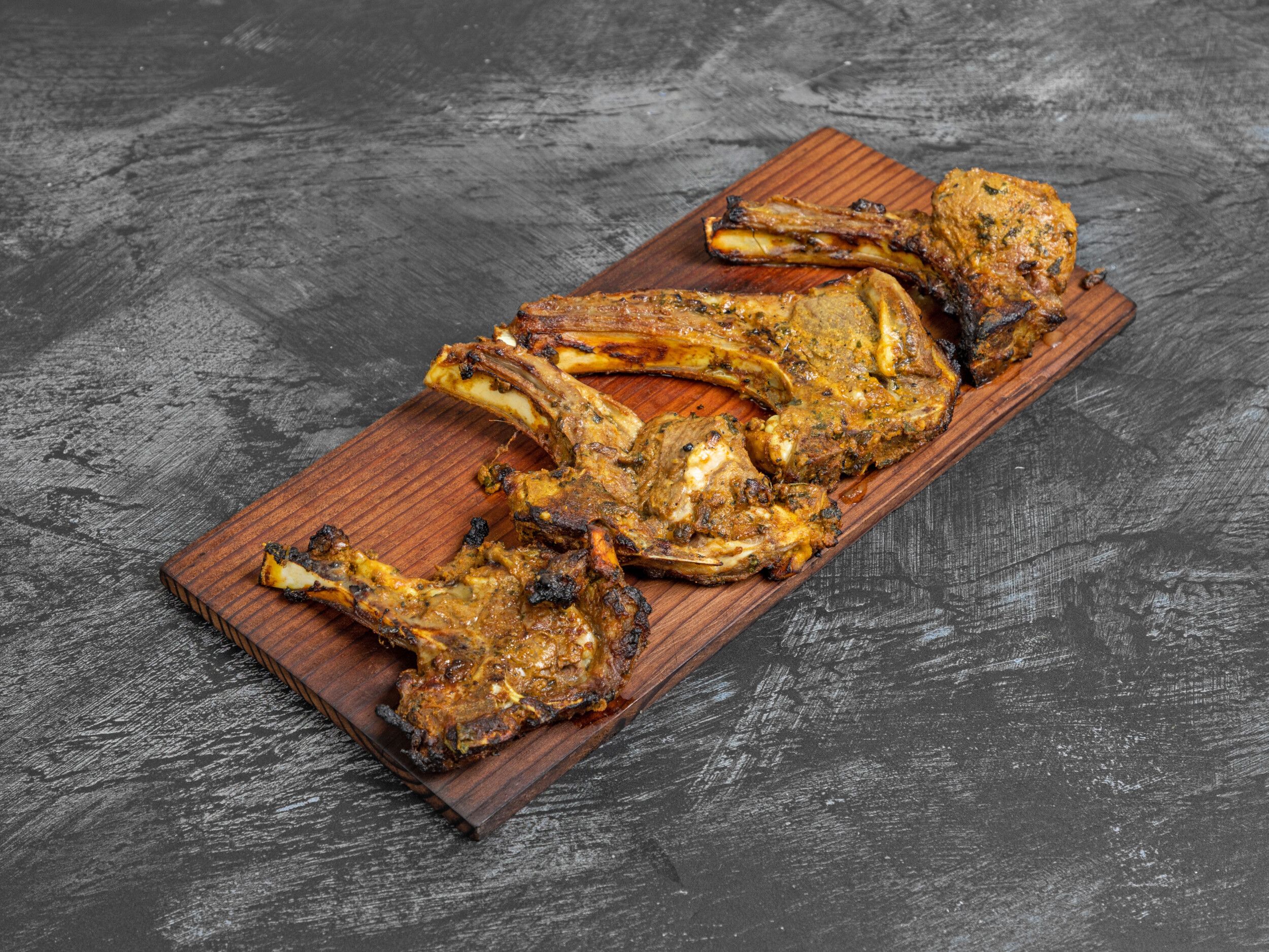 Grilled lamb chops seasoned with spices, placed on a dark wooden serving board over a dark grey surface.