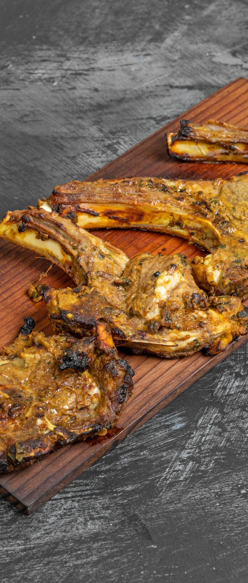 Grilled lamb chops seasoned with spices, placed on a dark wooden serving board over a dark grey surface.
