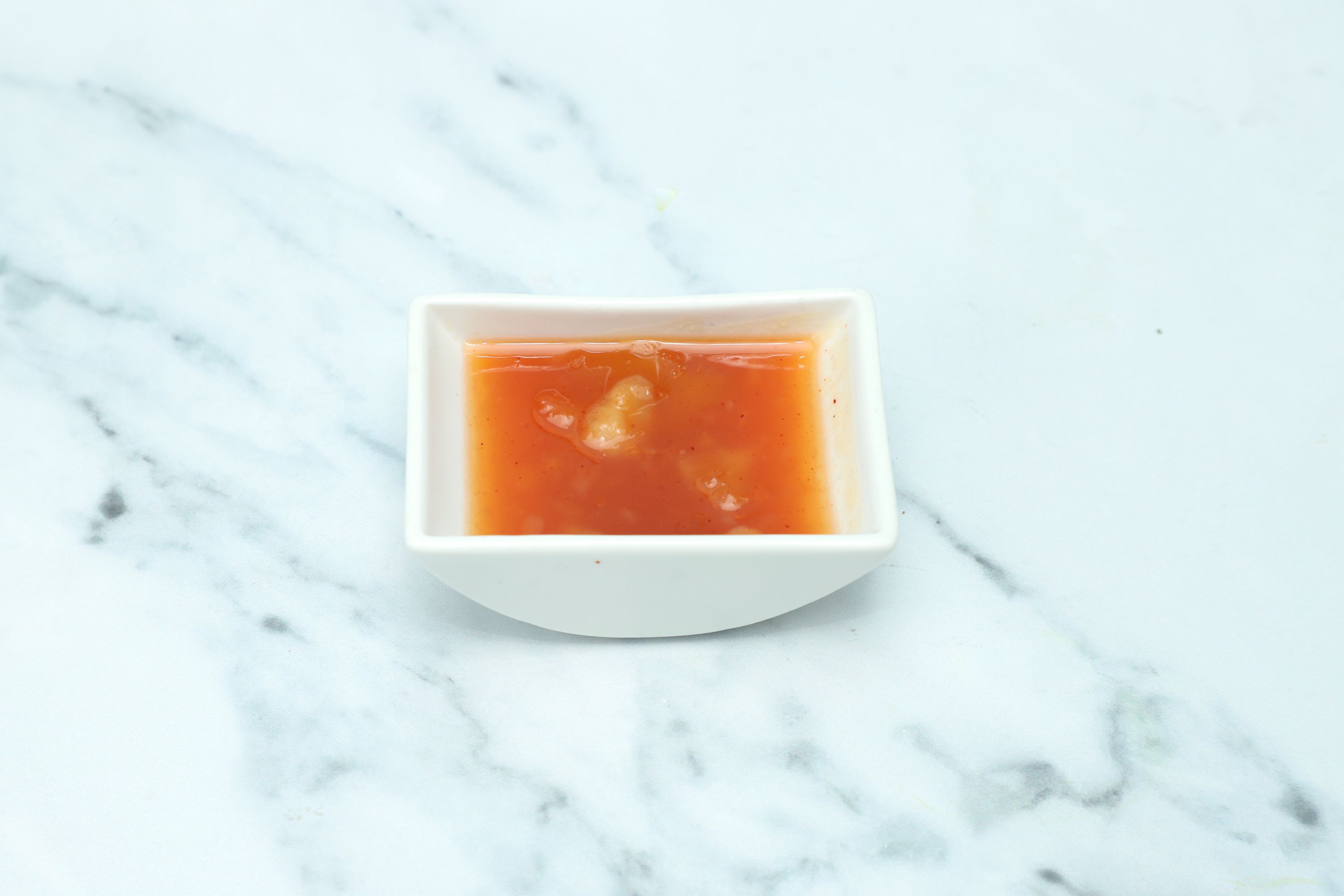 Small white square dish filled with vibrant orange-red sweet chili sauce on a marble surface.