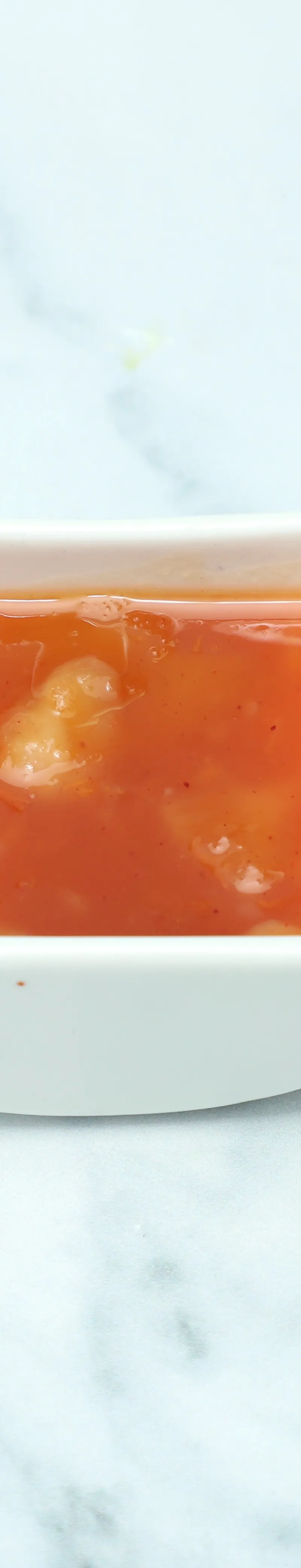 Small white square dish filled with vibrant orange-red sweet chili sauce on a marble surface.
