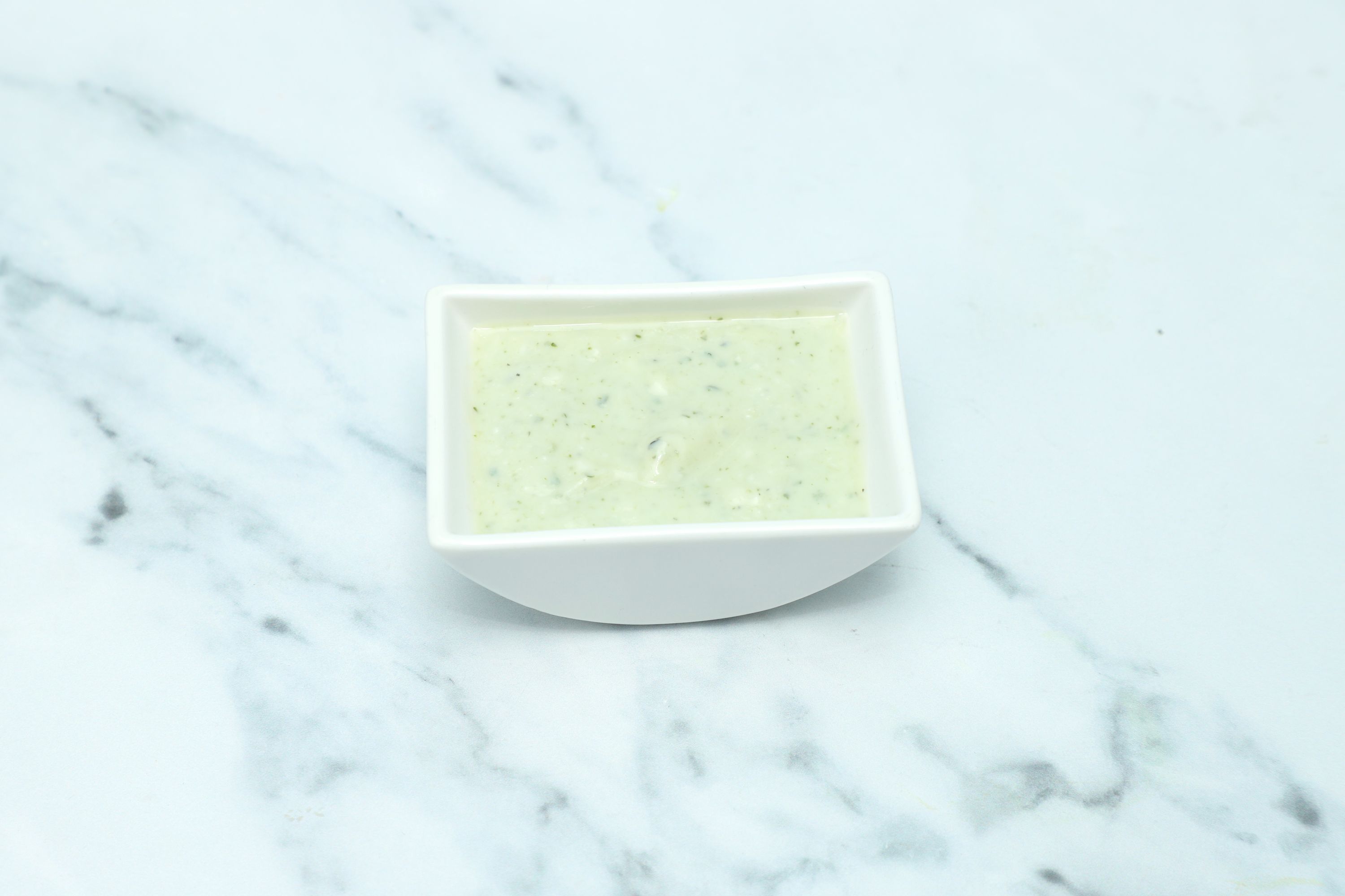 Small white square bowl filled with light green mint sauce sits on a marble surface.