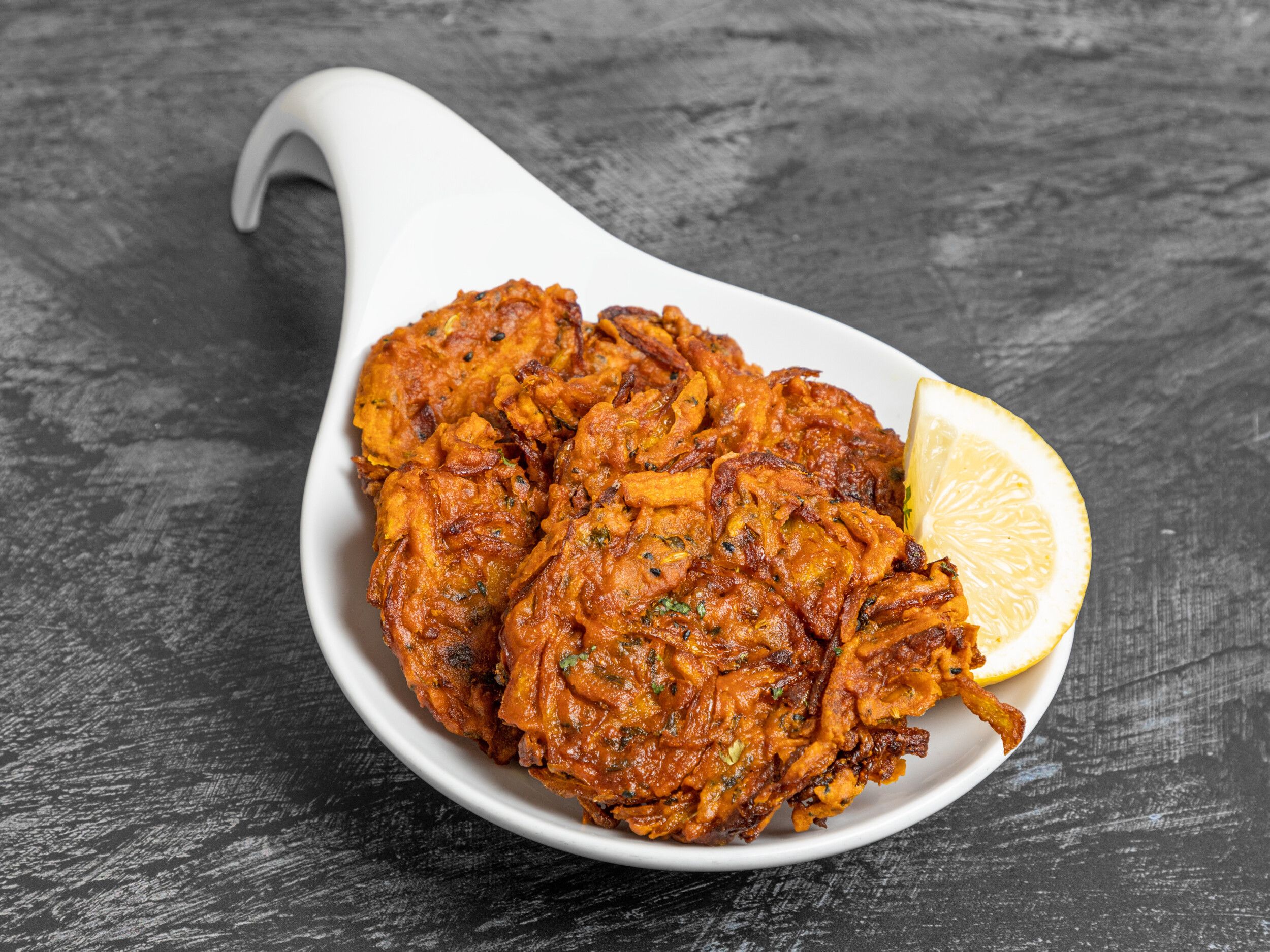Several golden brown onion bhajis with green herbs in a white serving dish with a lemon wedge