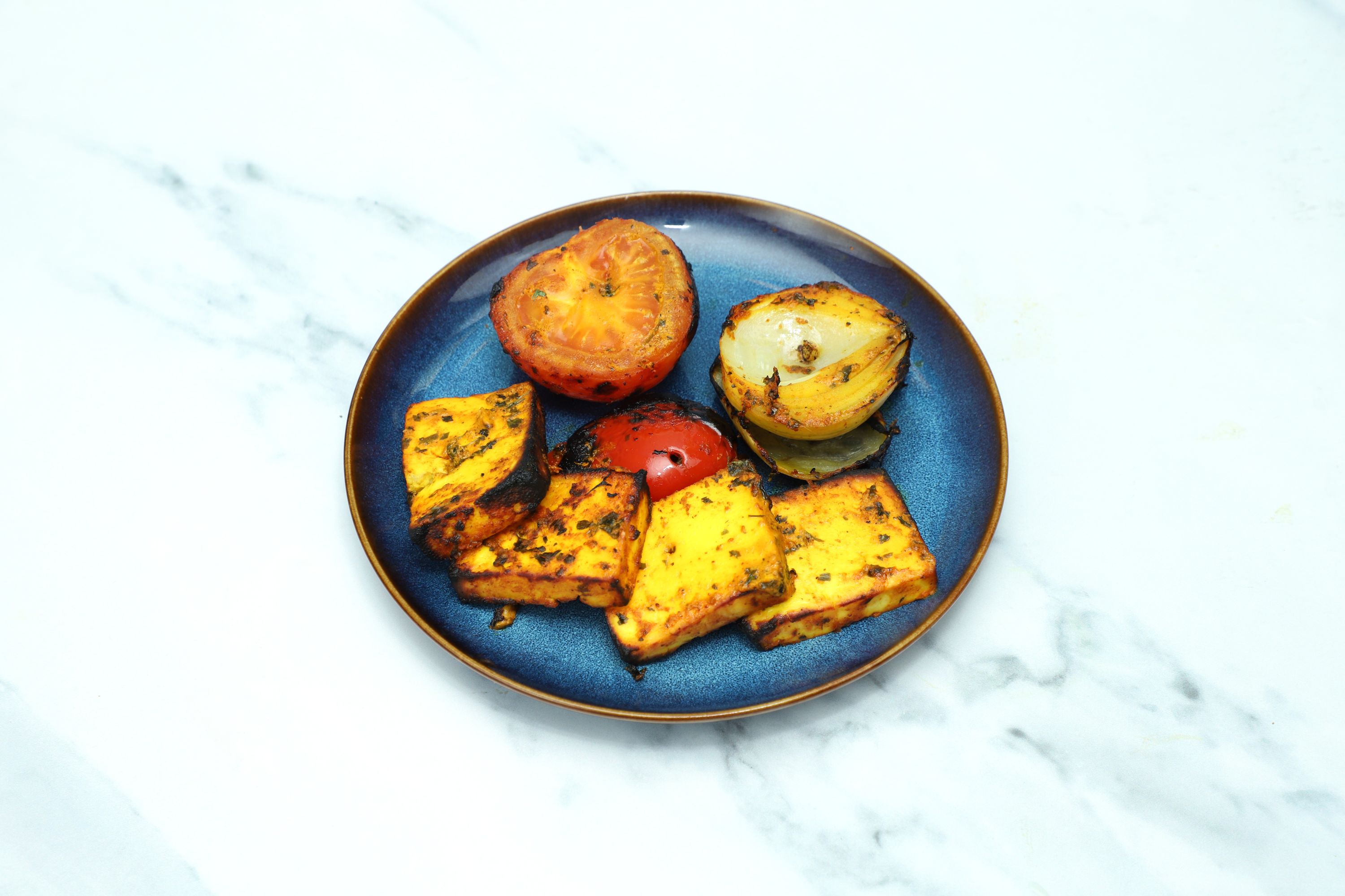 Grilled paneer cubes, two charred tomato halves, and a grilled onion on a blue plate.