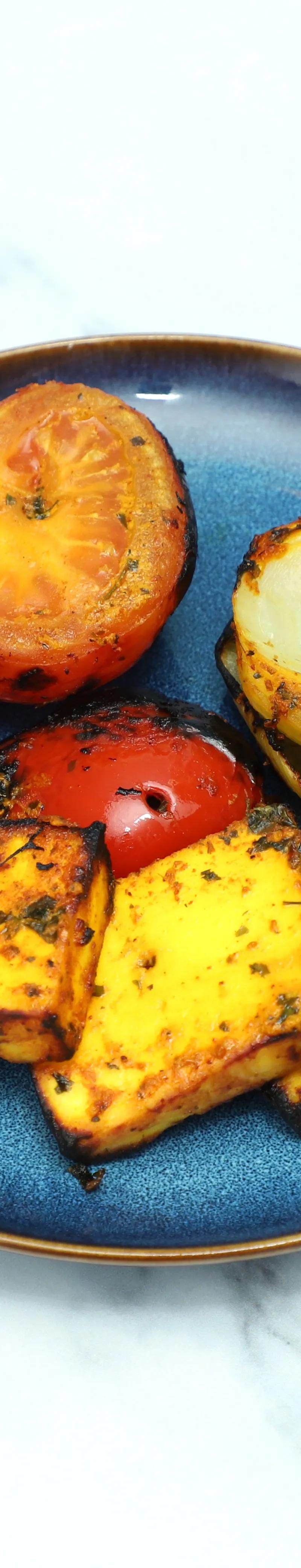 Grilled paneer cubes, two charred tomato halves, and a grilled onion on a blue plate.