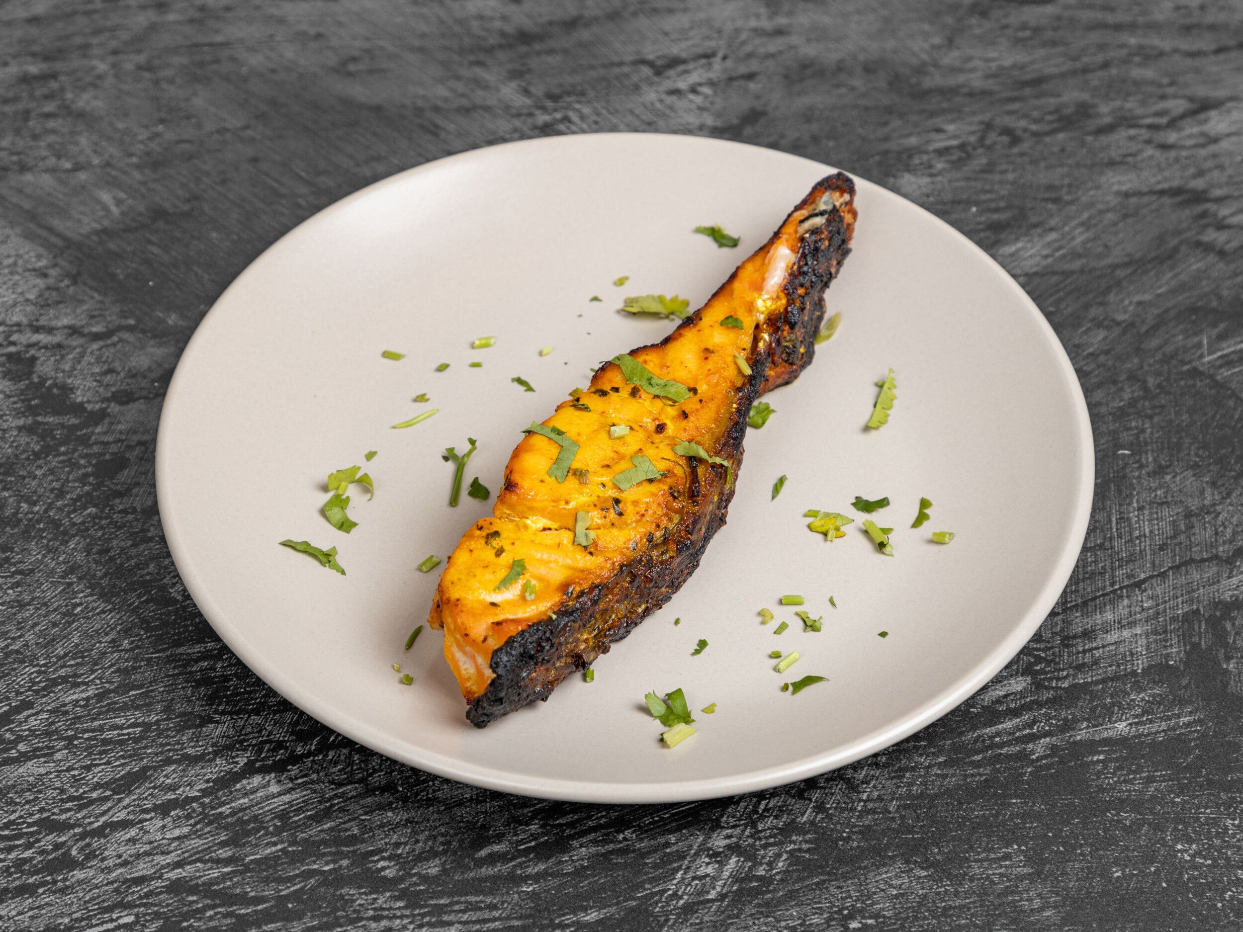 Tandoori salmon fillet garnished with chopped cilantro on a beige plate against a dark background.