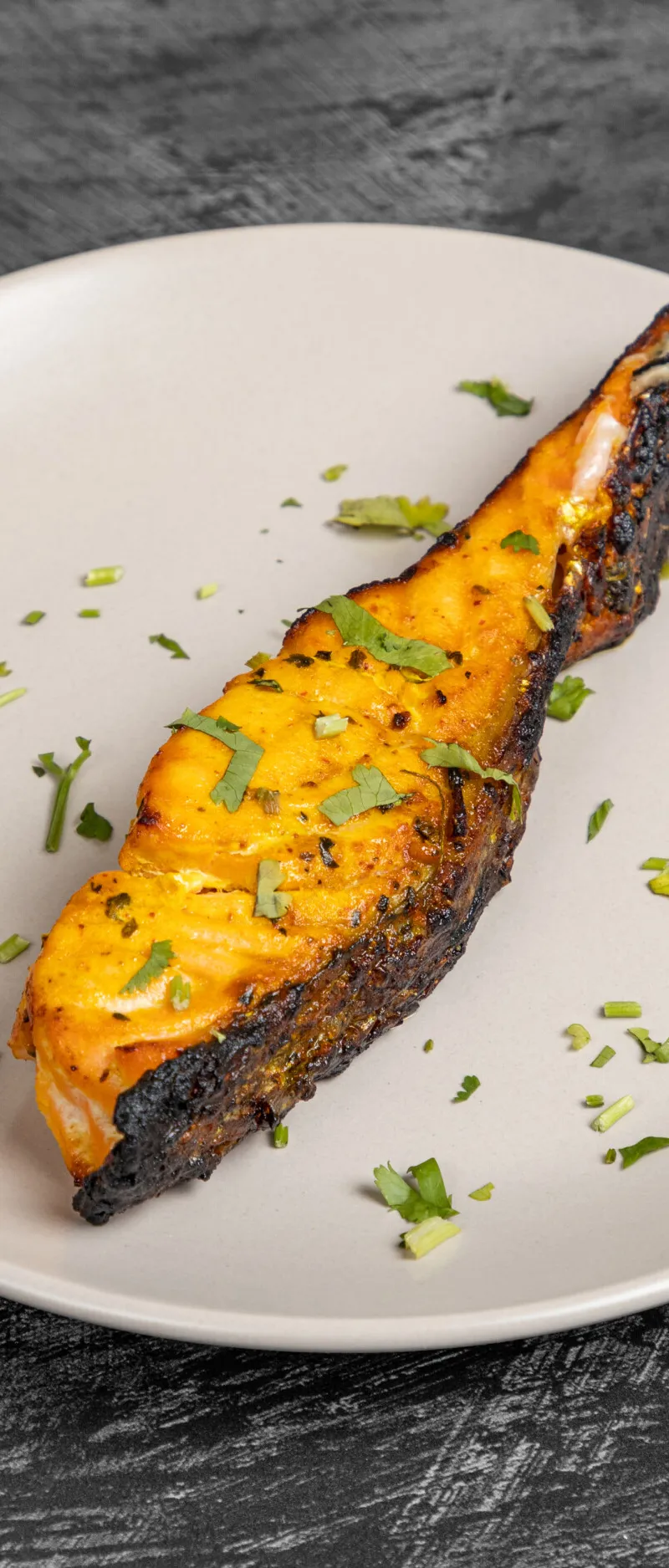 Tandoori salmon fillet garnished with chopped cilantro on a beige plate against a dark background.