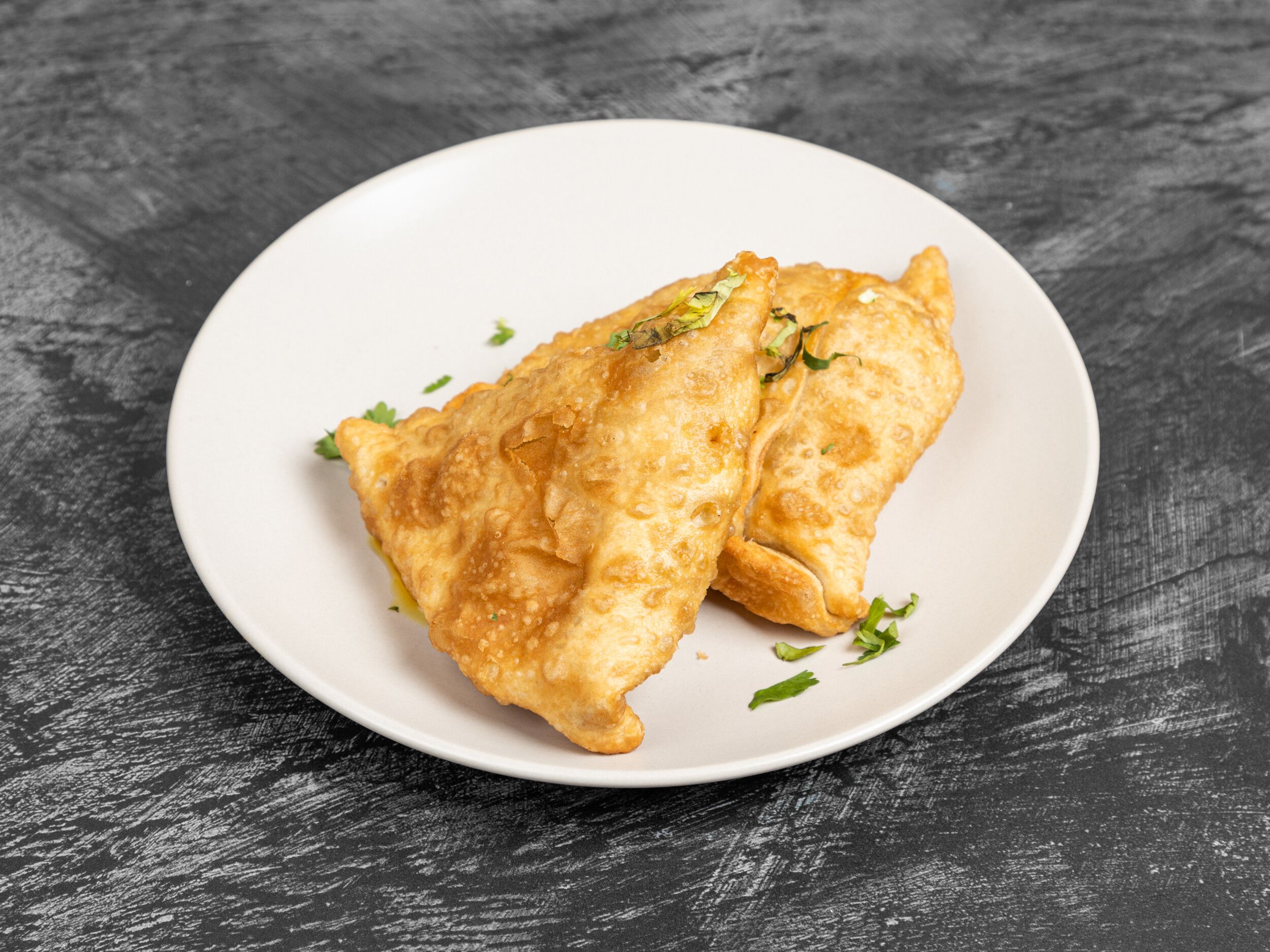 Two golden-brown fried samosas, triangular and flaky, garnished with fresh green herbs on a white plate.