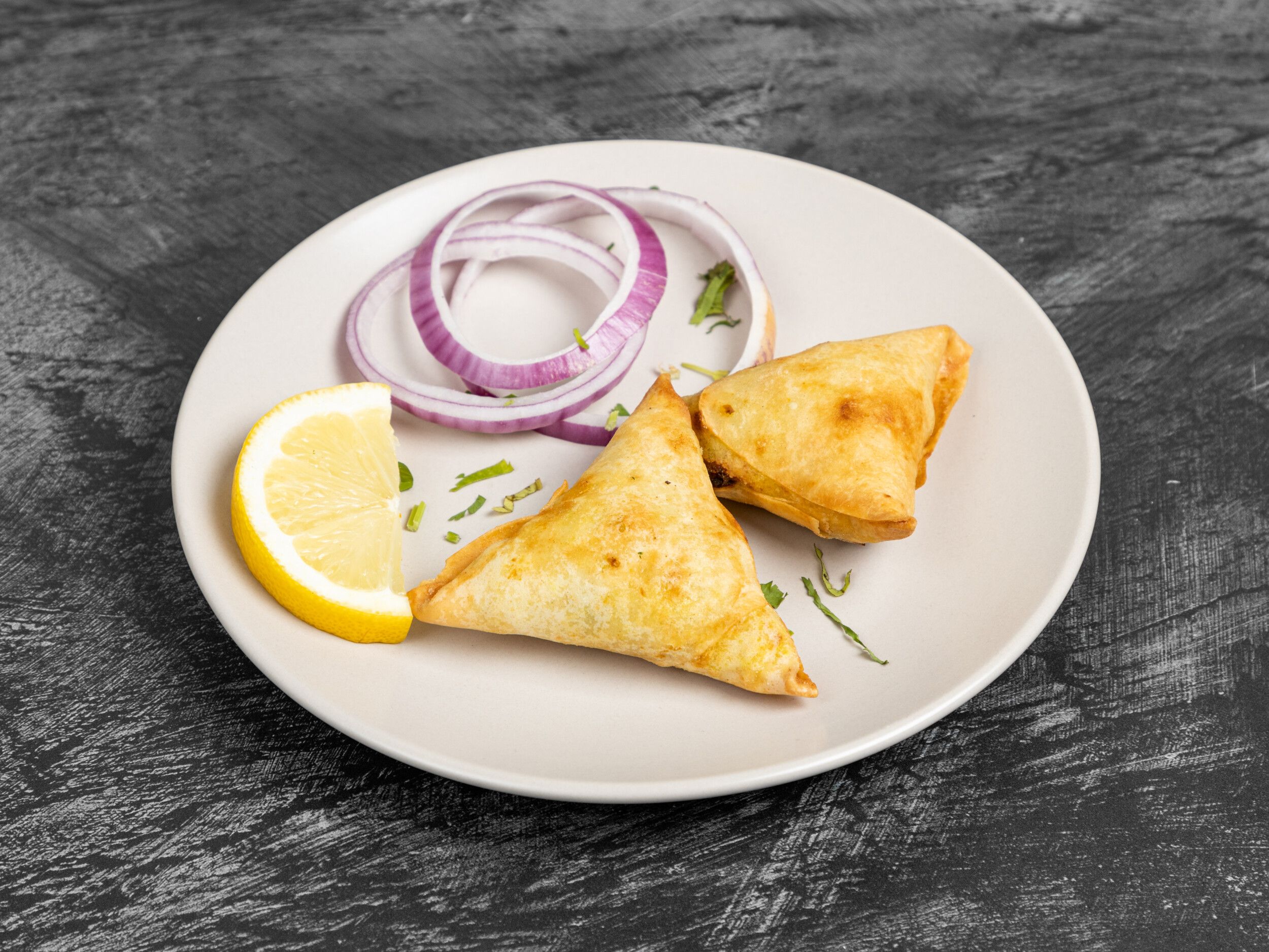 Two golden-brown samosas on a white plate with a lemon slice and red onion rings.
