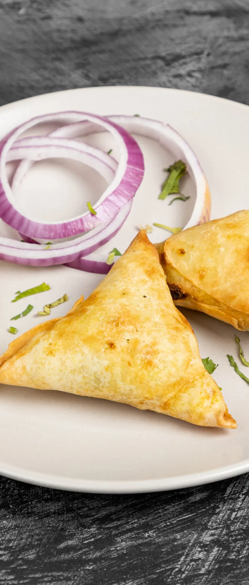 Two golden-brown samosas on a white plate with a lemon slice and red onion rings.