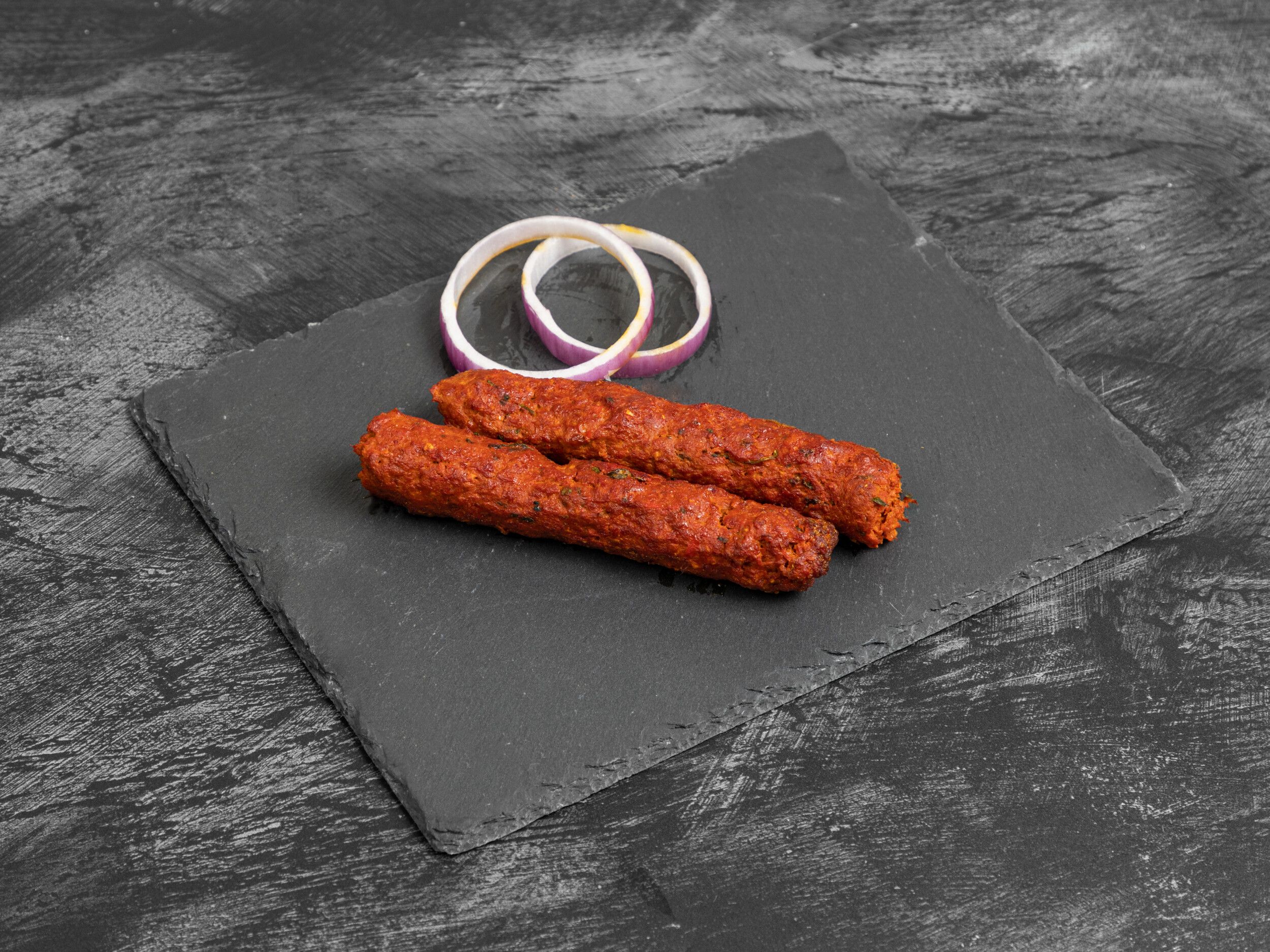 Two red seekh kebabs and two white and purple onion rings on a dark grey slate serving board.