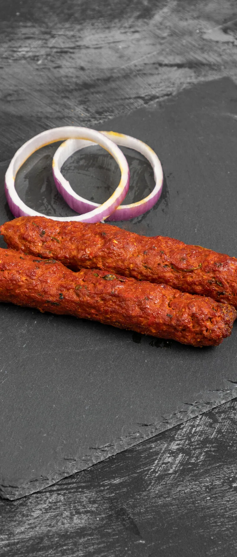 Two red seekh kebabs and two white and purple onion rings on a dark grey slate serving board.
