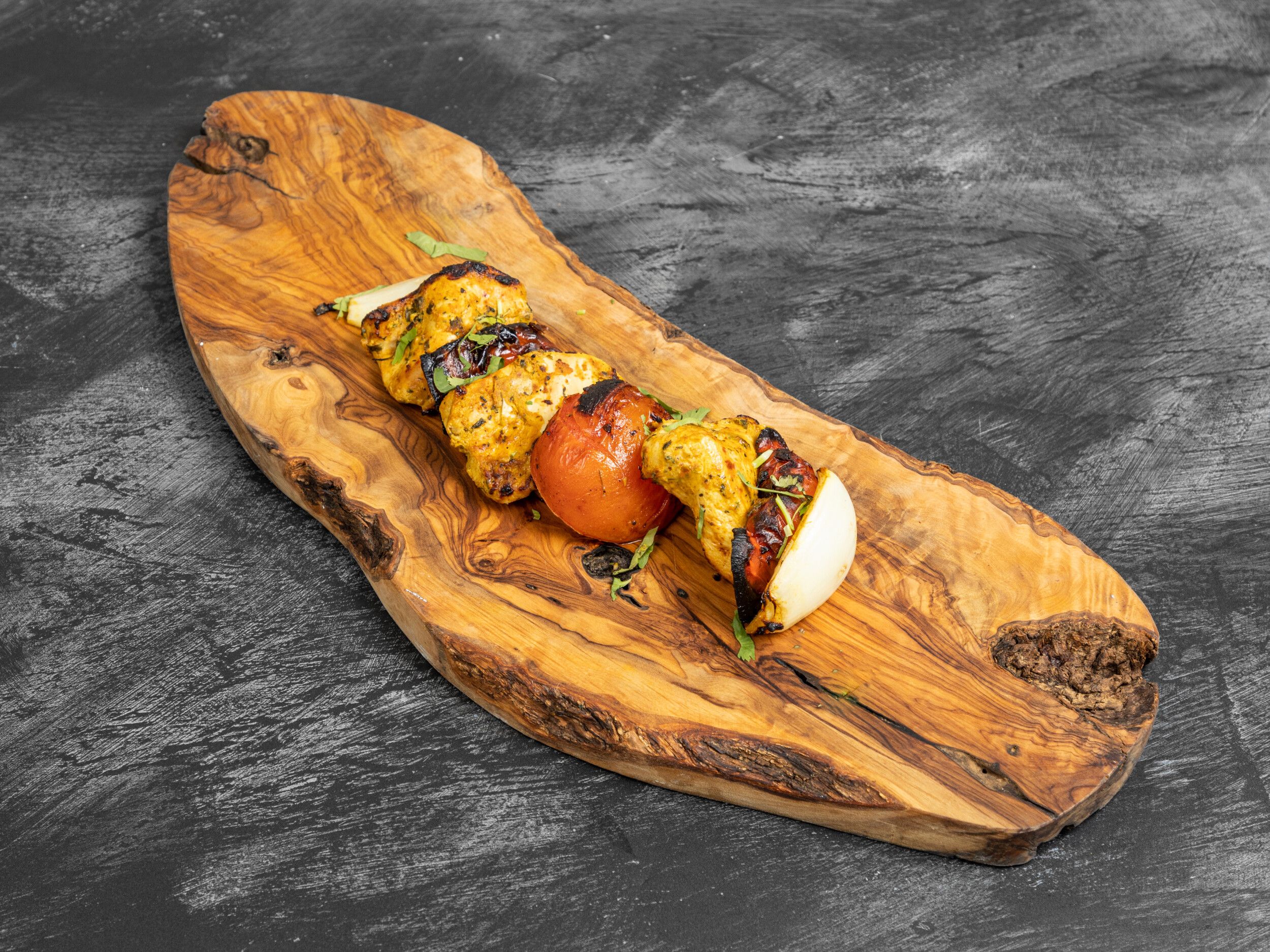 Grilled chicken pieces, tomatoes, and onions on a skewer, served on a rustic wooden board.