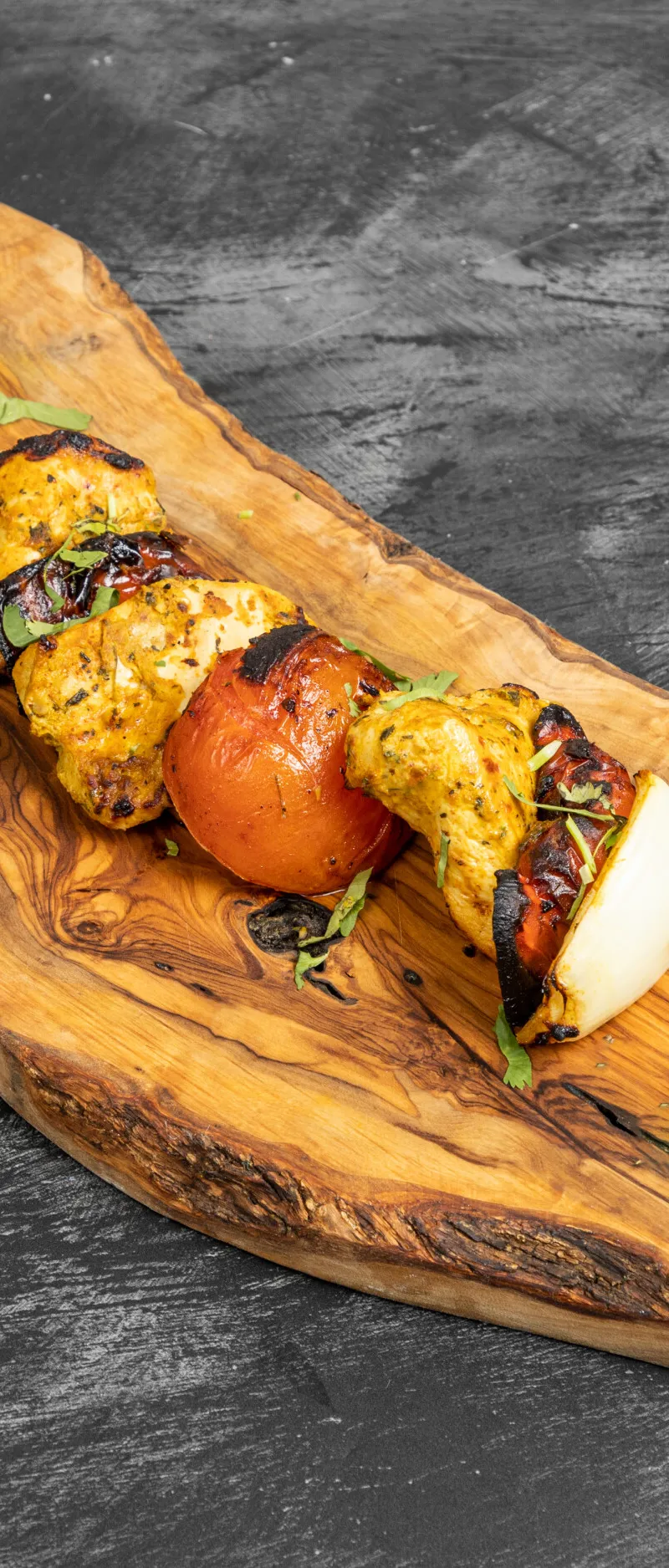 Grilled chicken pieces, tomatoes, and onions on a skewer, served on a rustic wooden board.