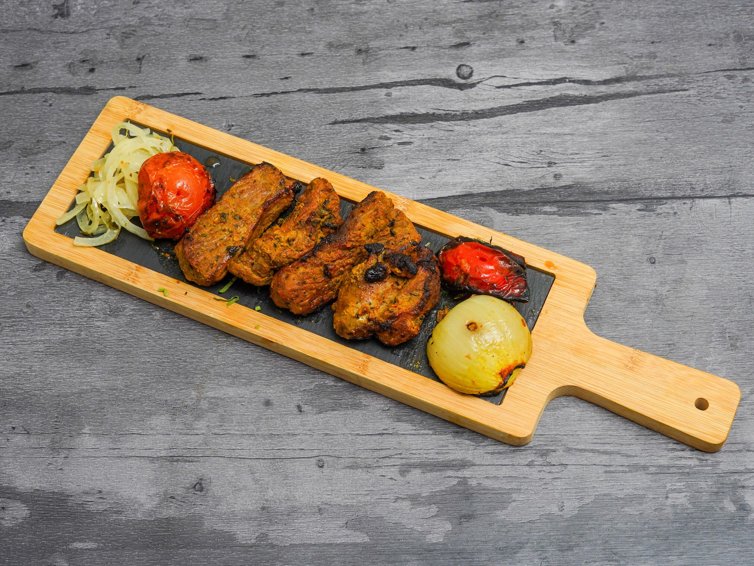 Grilled lamb chops, caramelized onions, and roasted tomatoes on a black slate platter with a wooden frame.