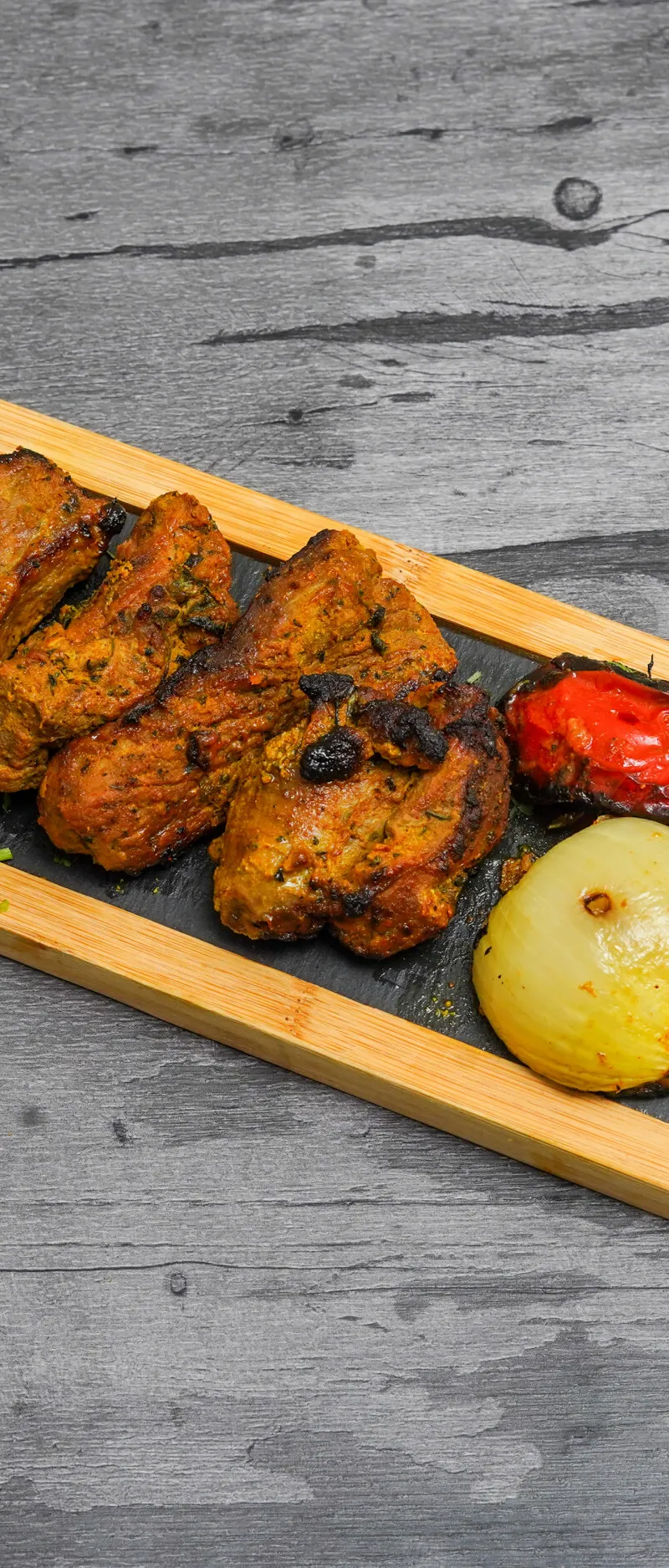 Grilled lamb chops, caramelized onions, and roasted tomatoes on a black slate platter with a wooden frame.