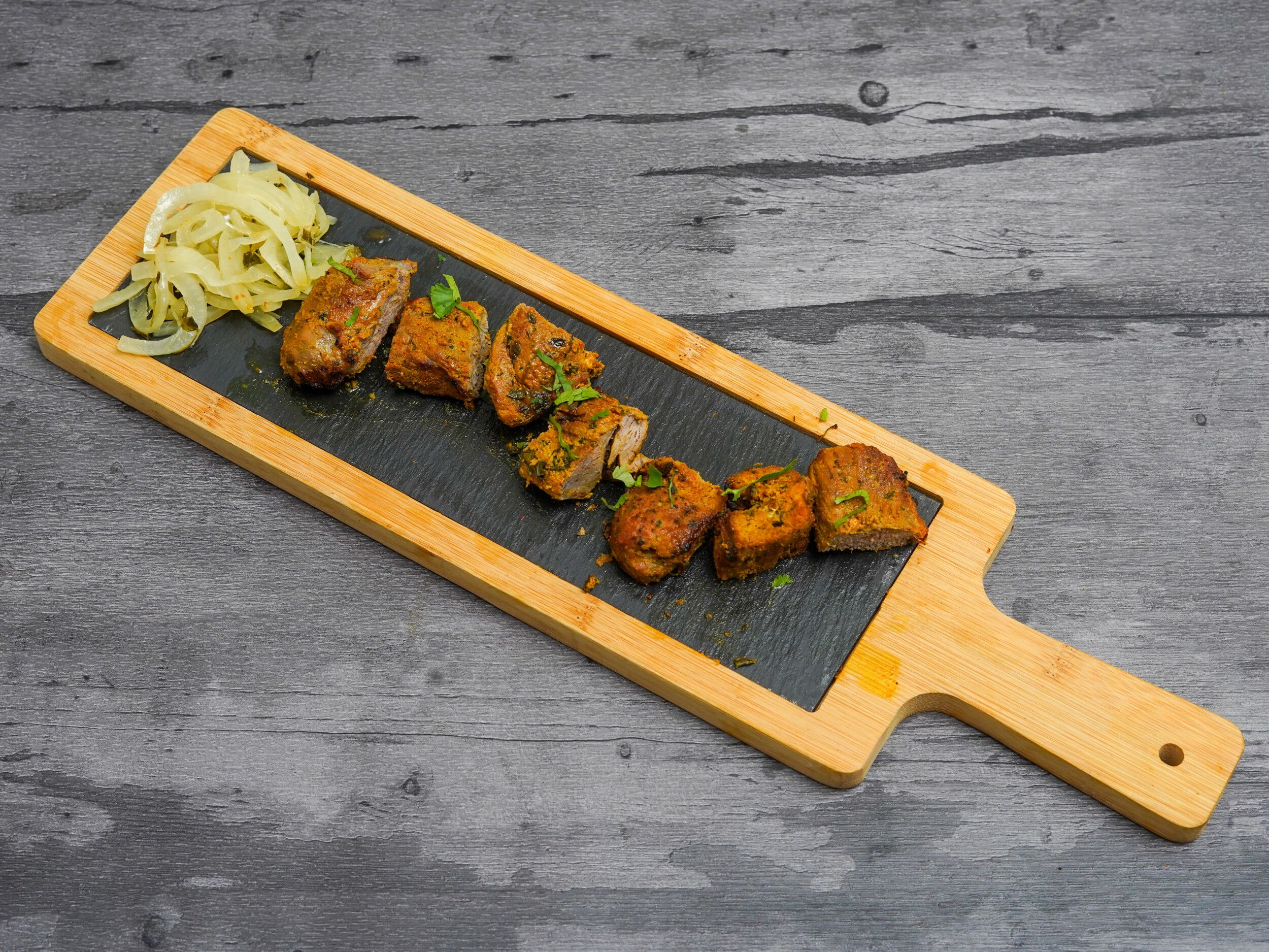 Lamb tikka pieces and sliced white onions on a black slate serving board with wooden trim and handle.