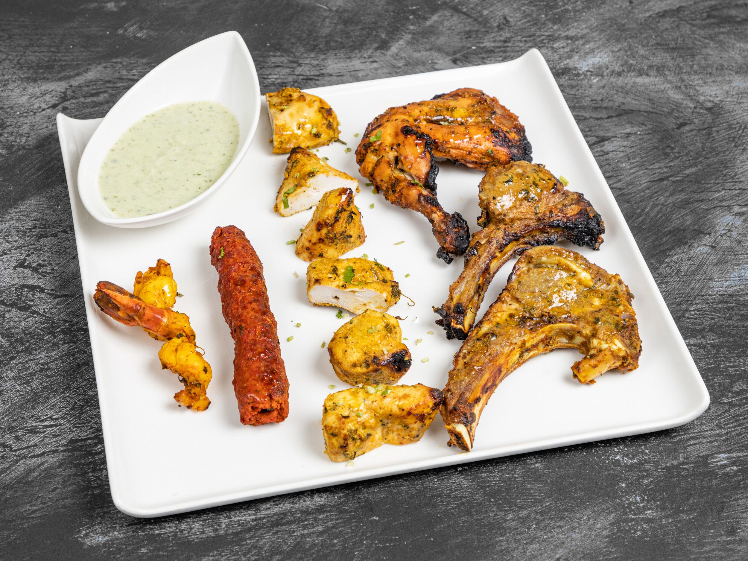 Grilled chicken drumstick, shrimp, lamb chops, seekh kebab, and chicken tikka on a white rectangular plate with green sauce.