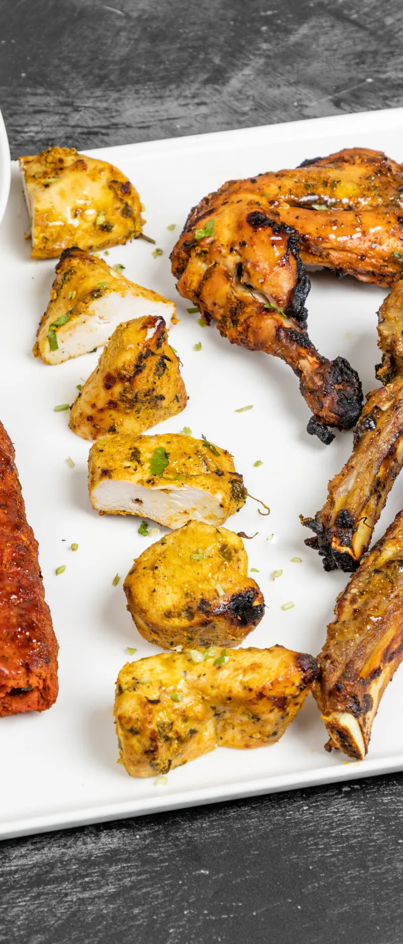 Grilled chicken drumstick, shrimp, lamb chops, seekh kebab, and chicken tikka on a white rectangular plate with green sauce.