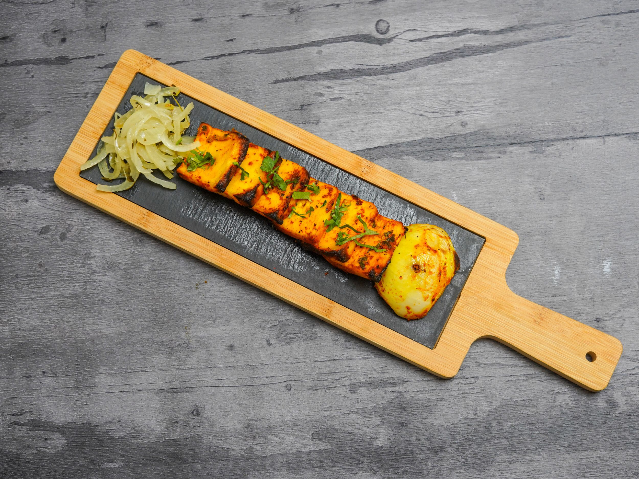 Golden paneer tikka skewers with charred edges, garnished with coriander, served with sliced onions and a grilled onion half on a black slate inlay paddle