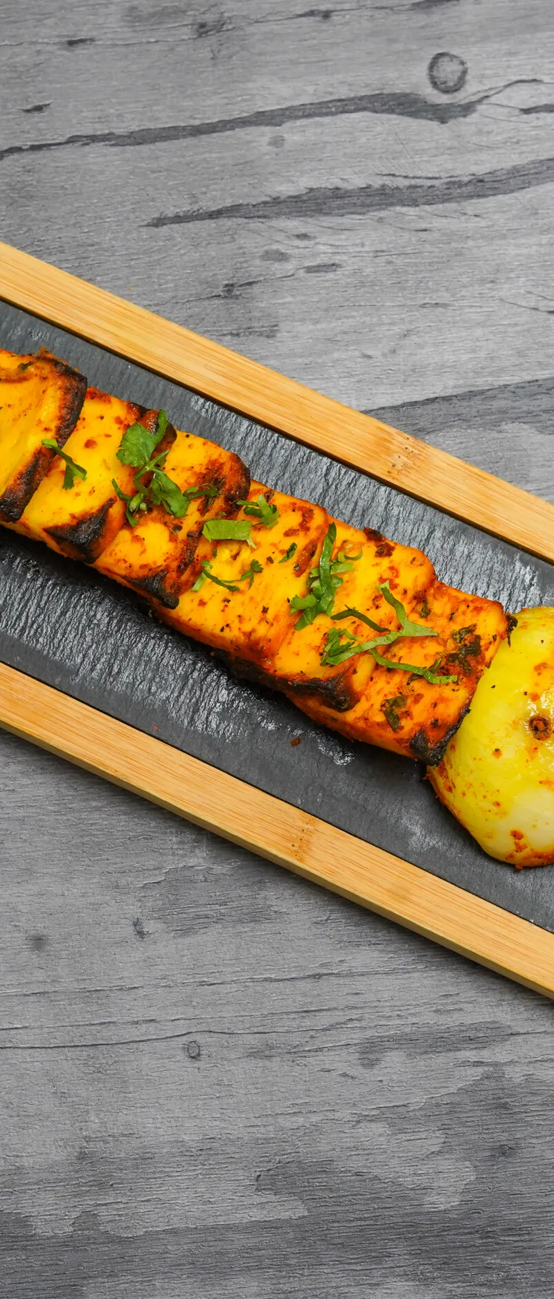 Golden paneer tikka skewers with charred edges, garnished with coriander, served with sliced onions and a grilled onion half on a black slate inlay paddle