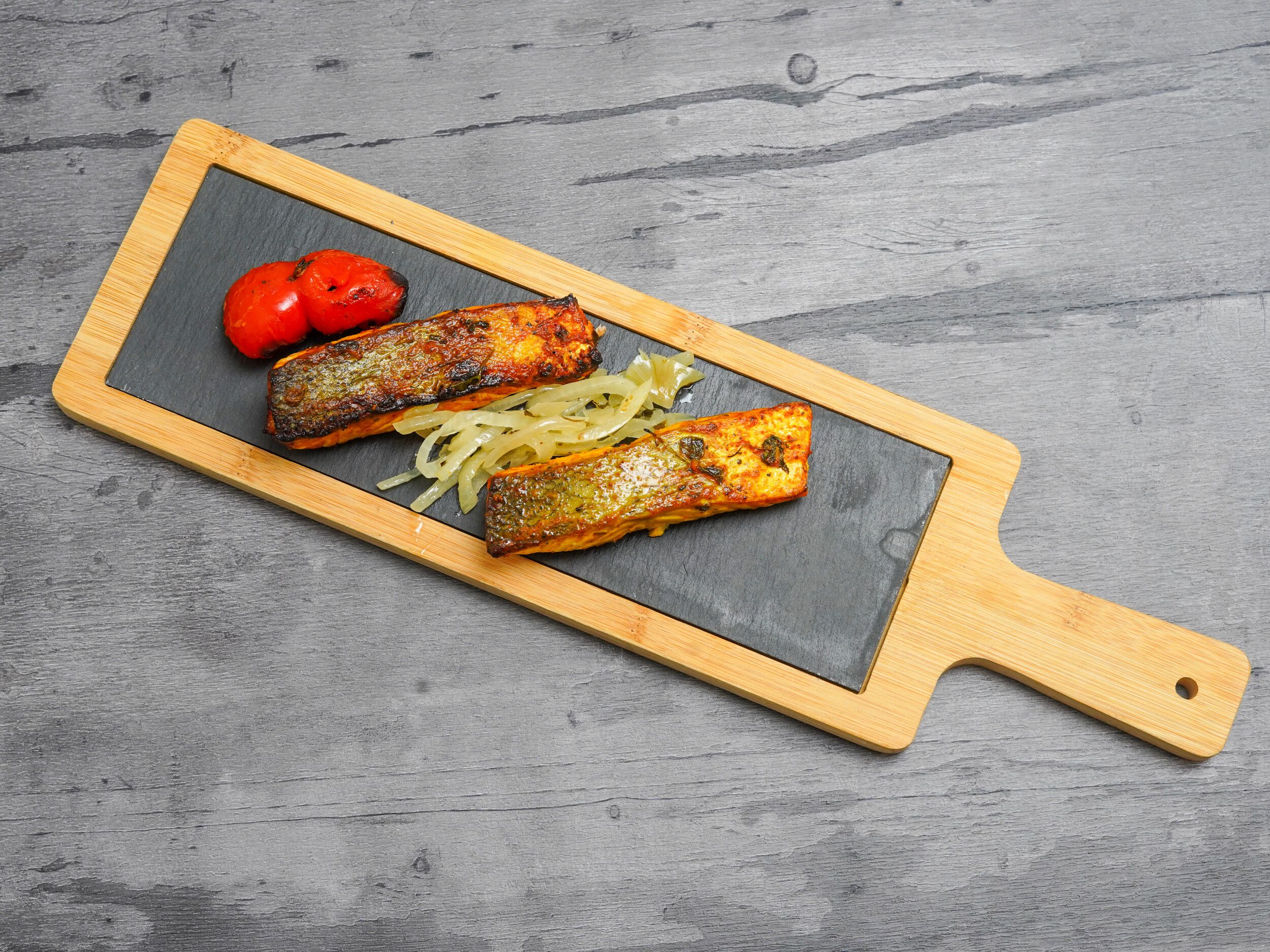 Two Tandoori salmon fillets, charred tomatoes, and caramelised onions on a black slate, set in a wooden tray.
