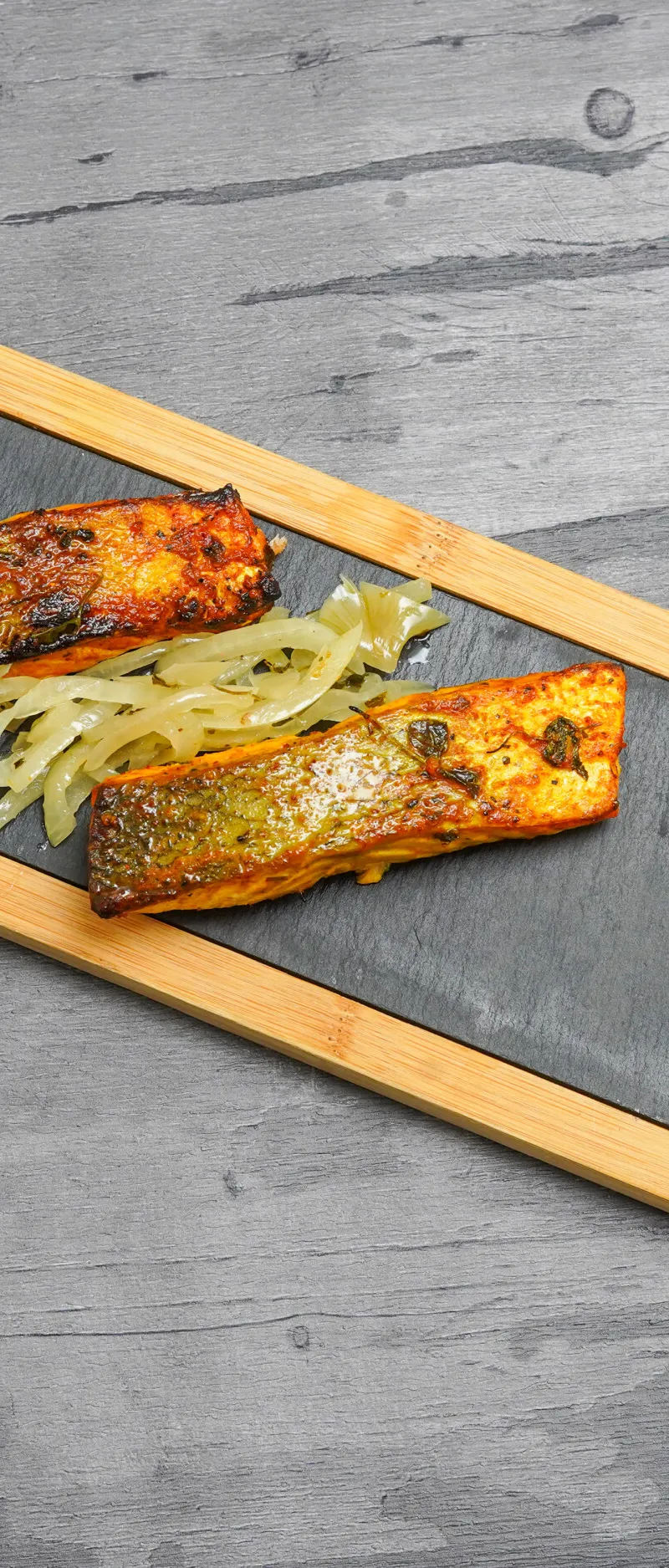 Two Tandoori salmon fillets, charred tomatoes, and caramelised onions on a black slate, set in a wooden tray.