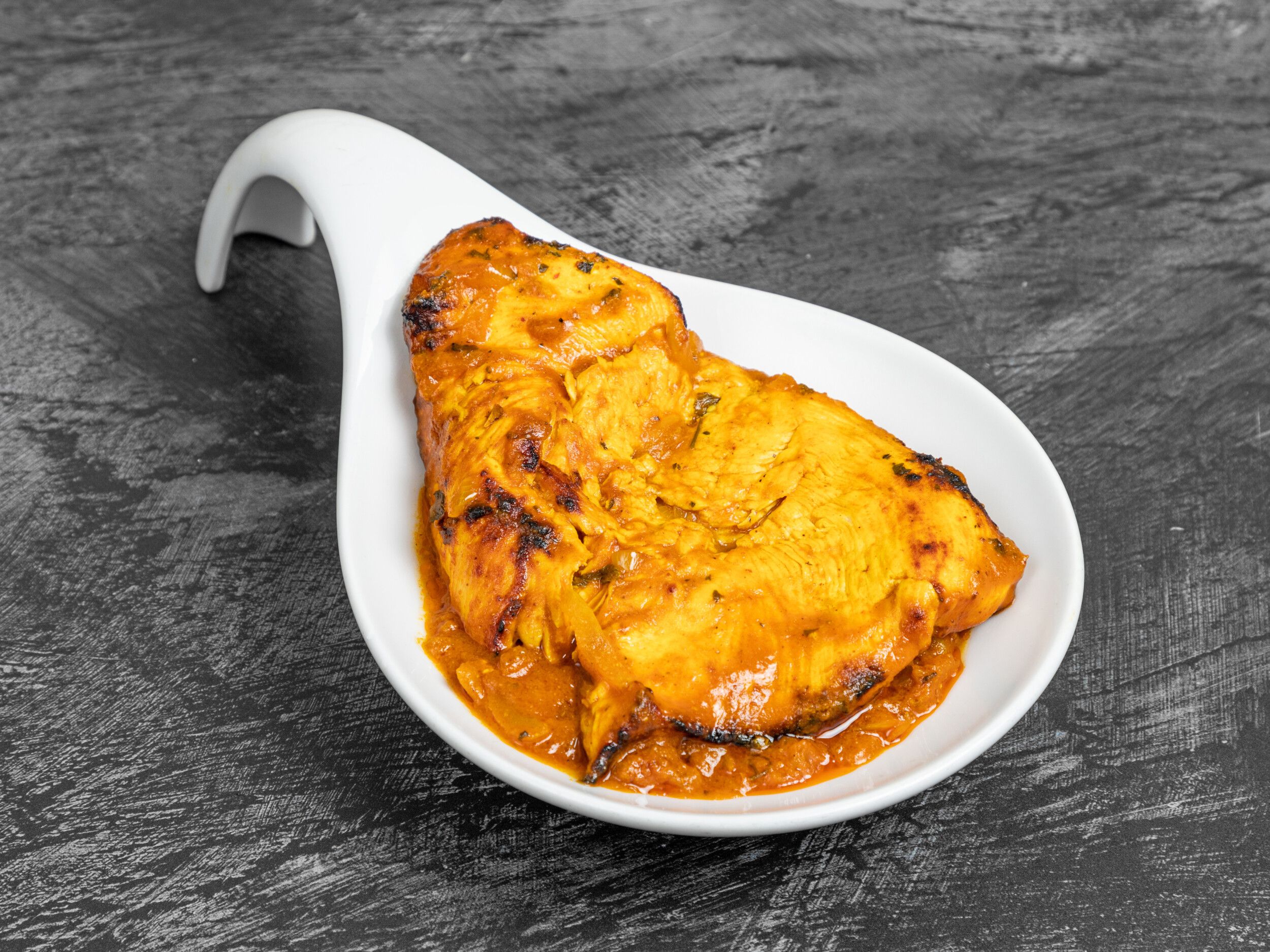 Bright orange tandoori chicken fillet with some charring, served in white modern spoon-shaped dish.