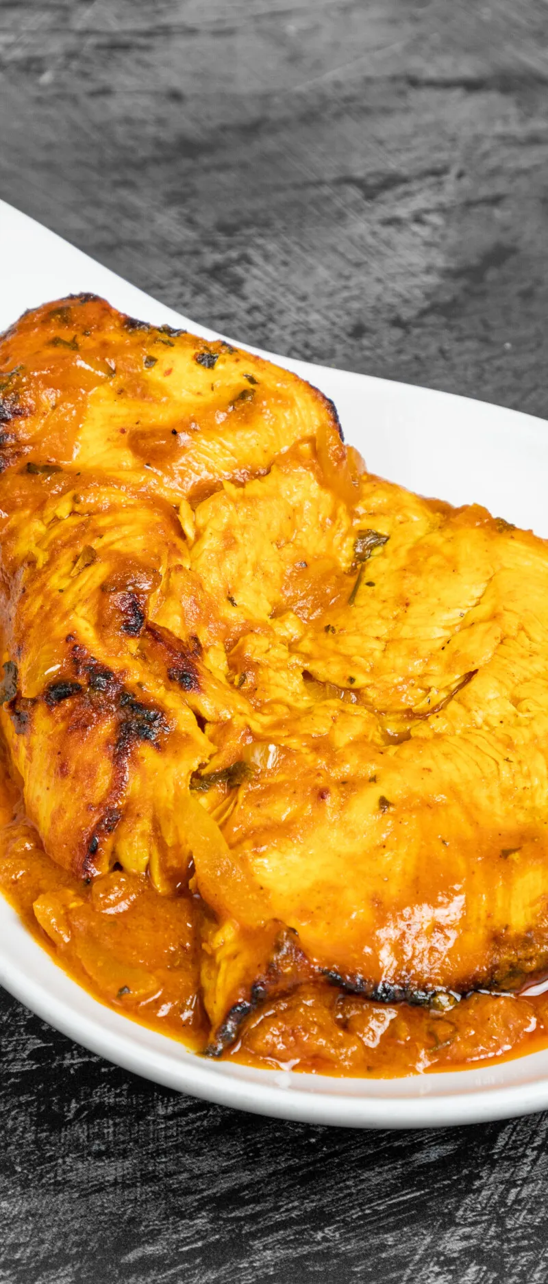 Bright orange tandoori chicken fillet with some charring, served in white modern spoon-shaped dish.