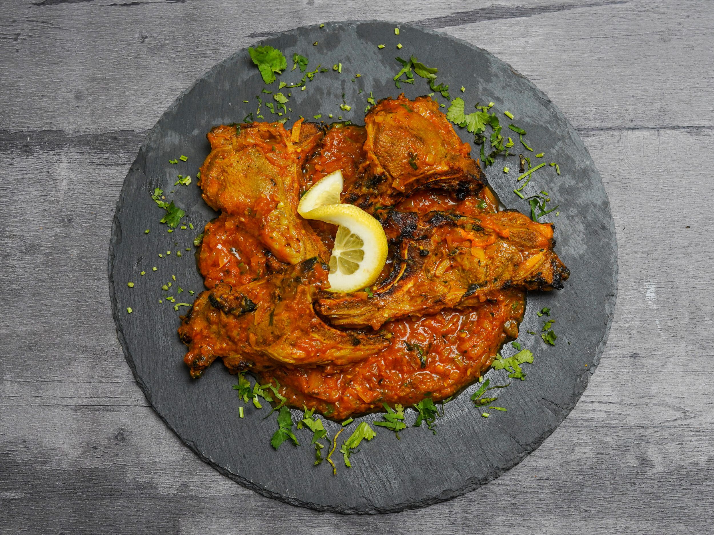 Grilled lamb chops in rich orange sauce on a dark slate platter, garnished with cilantro and lemon