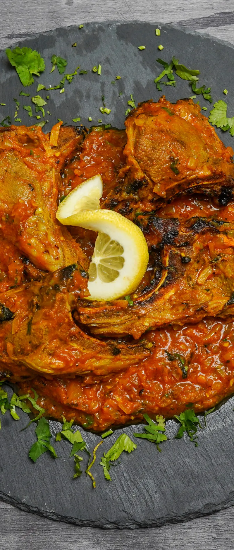 Grilled lamb chops in rich orange sauce on a dark slate platter, garnished with cilantro and lemon