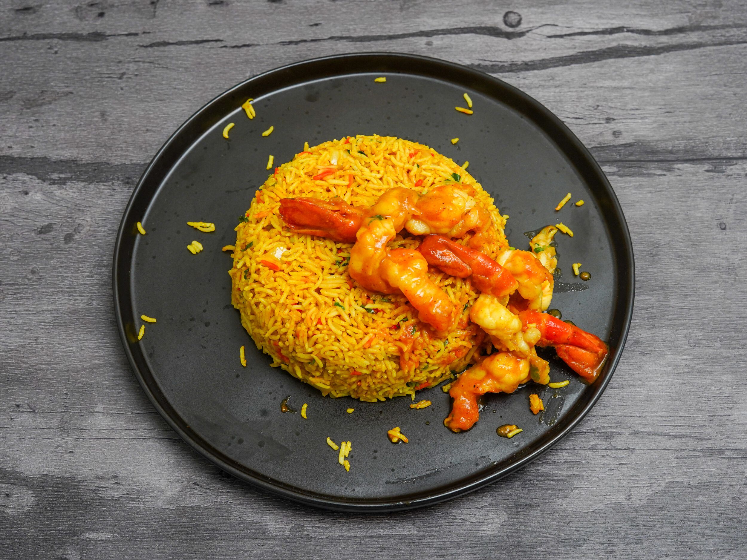 Bright yellow king prawn biryani in a black dish on a dark wooden surface, garnished with chili.